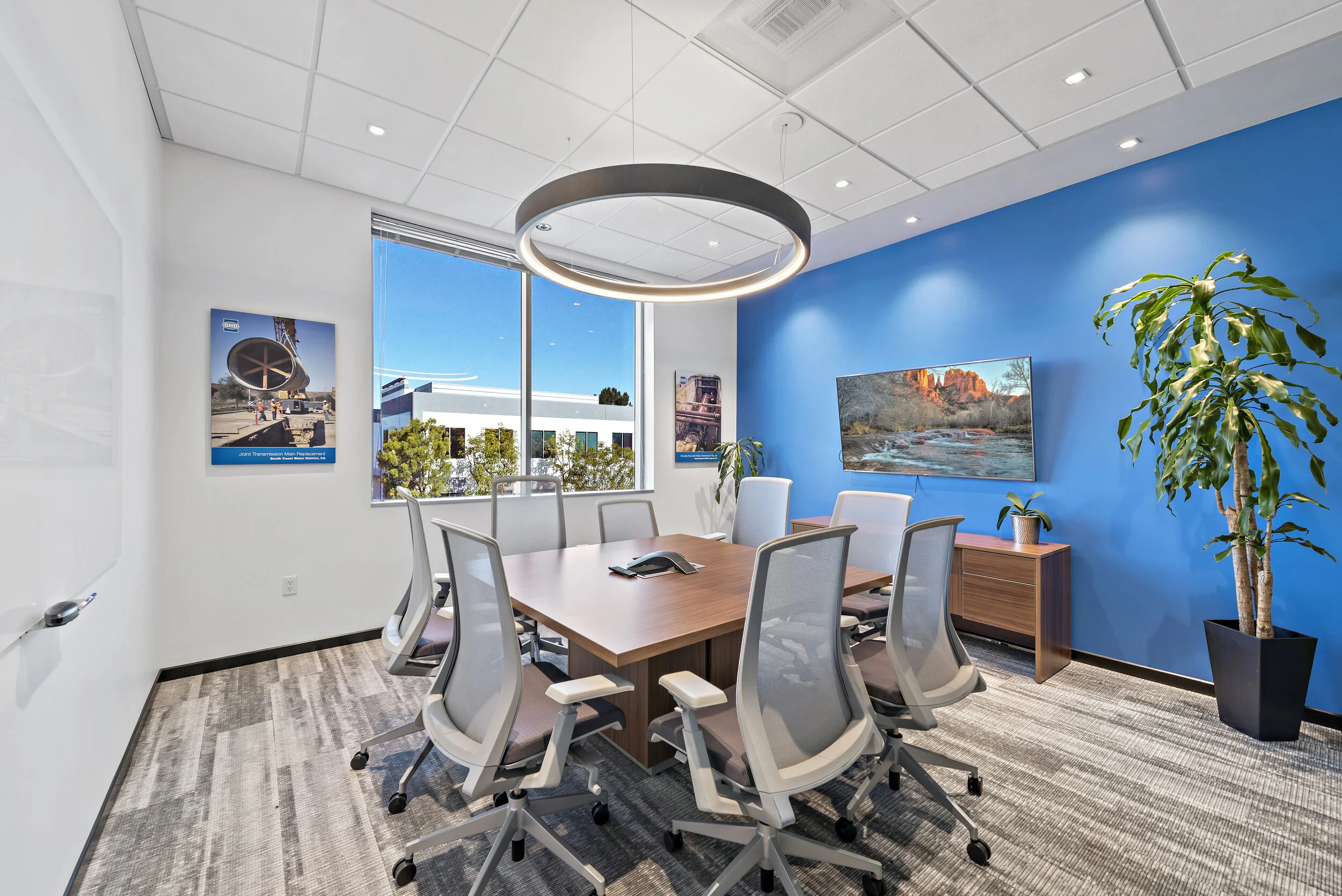 Modern conference room with white and blue walls, a large wooden table surrounded by nine white office chairs, a flat-screen TV on the blue wall, large window with cityscape view, potted plants, and a circular ceiling light fixture.