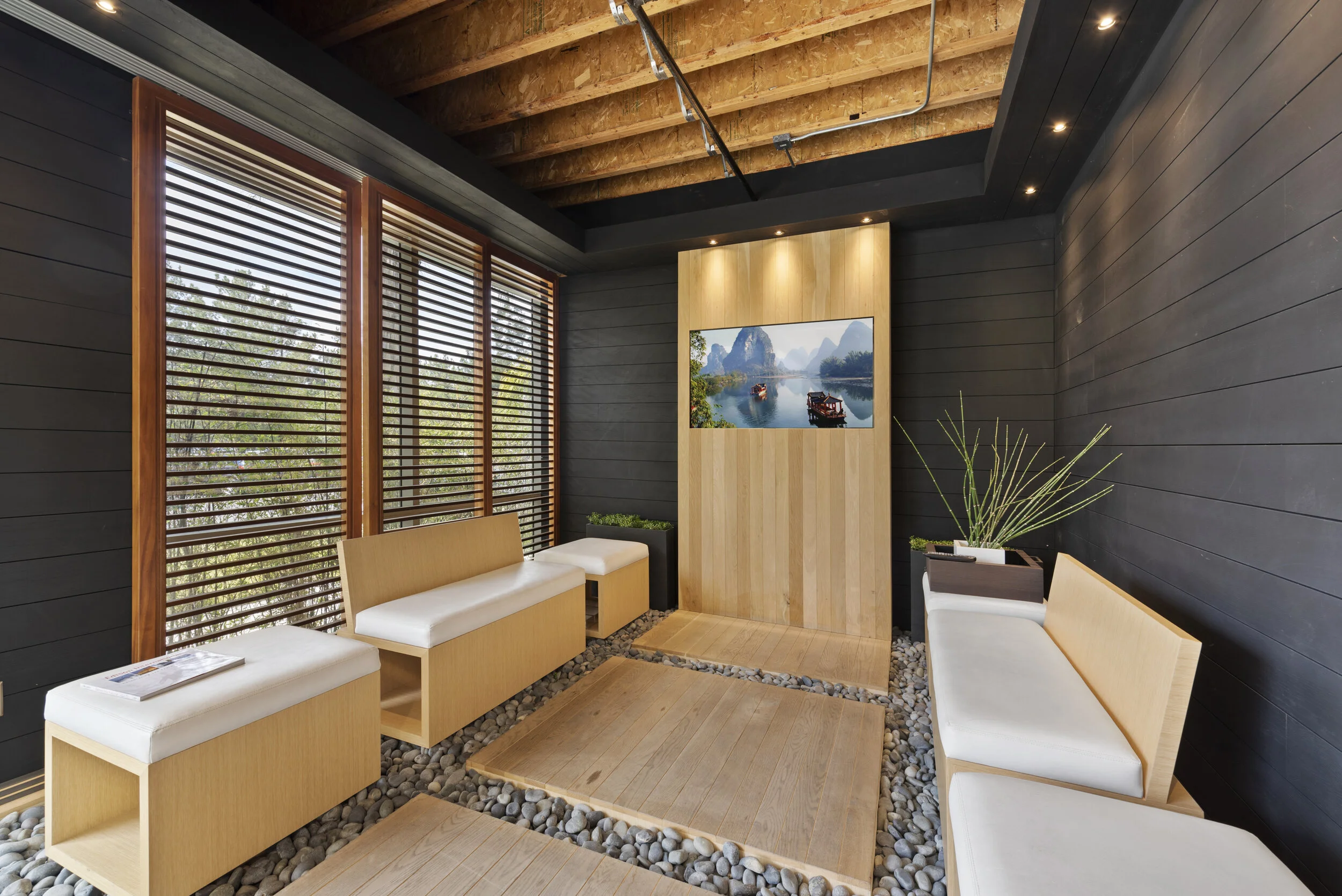 Modern waiting or lounge area with black wood-paneled walls, wooden benches with white cushions, a large window with wooden blinds, and a flat-screen TV displaying a river scene with boats and mountains.