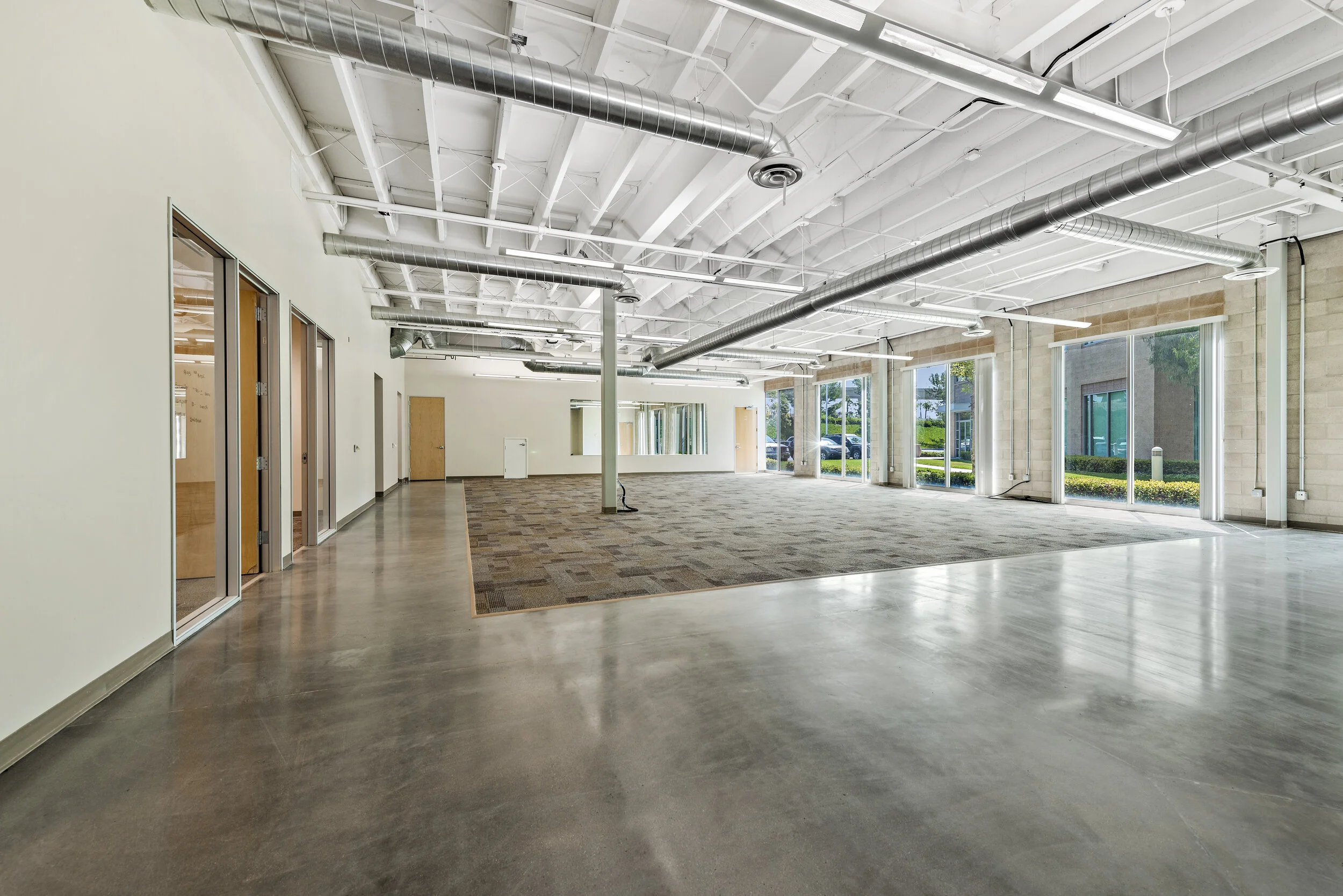 Empty commercial office space with large windows, polished concrete floors, exposed ductwork, and a corner area with carpet.