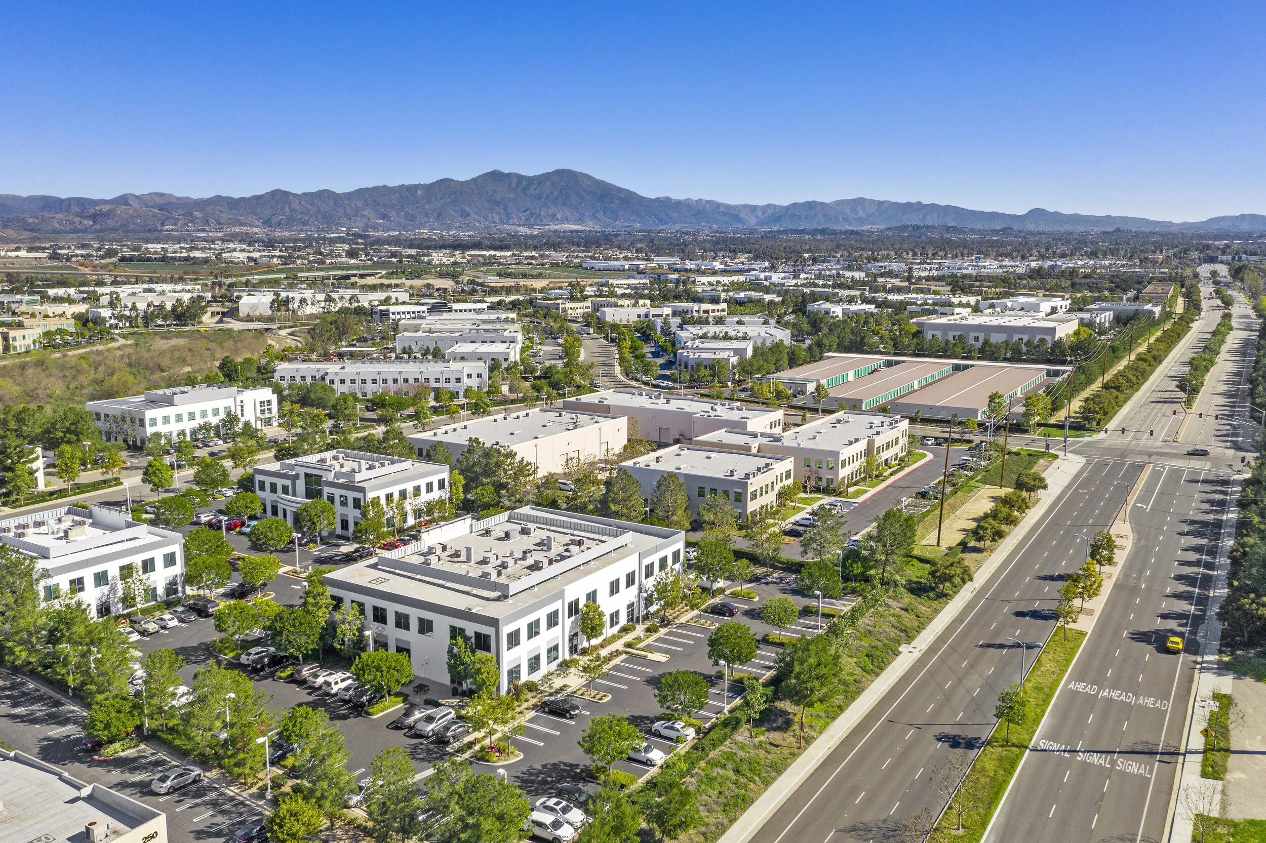 Aerial view of a commercial area with modern white office buildings, parking lots, green trees, and a wide road with street signs and light poles, set against a backdrop of mountains under a clear blue sky.