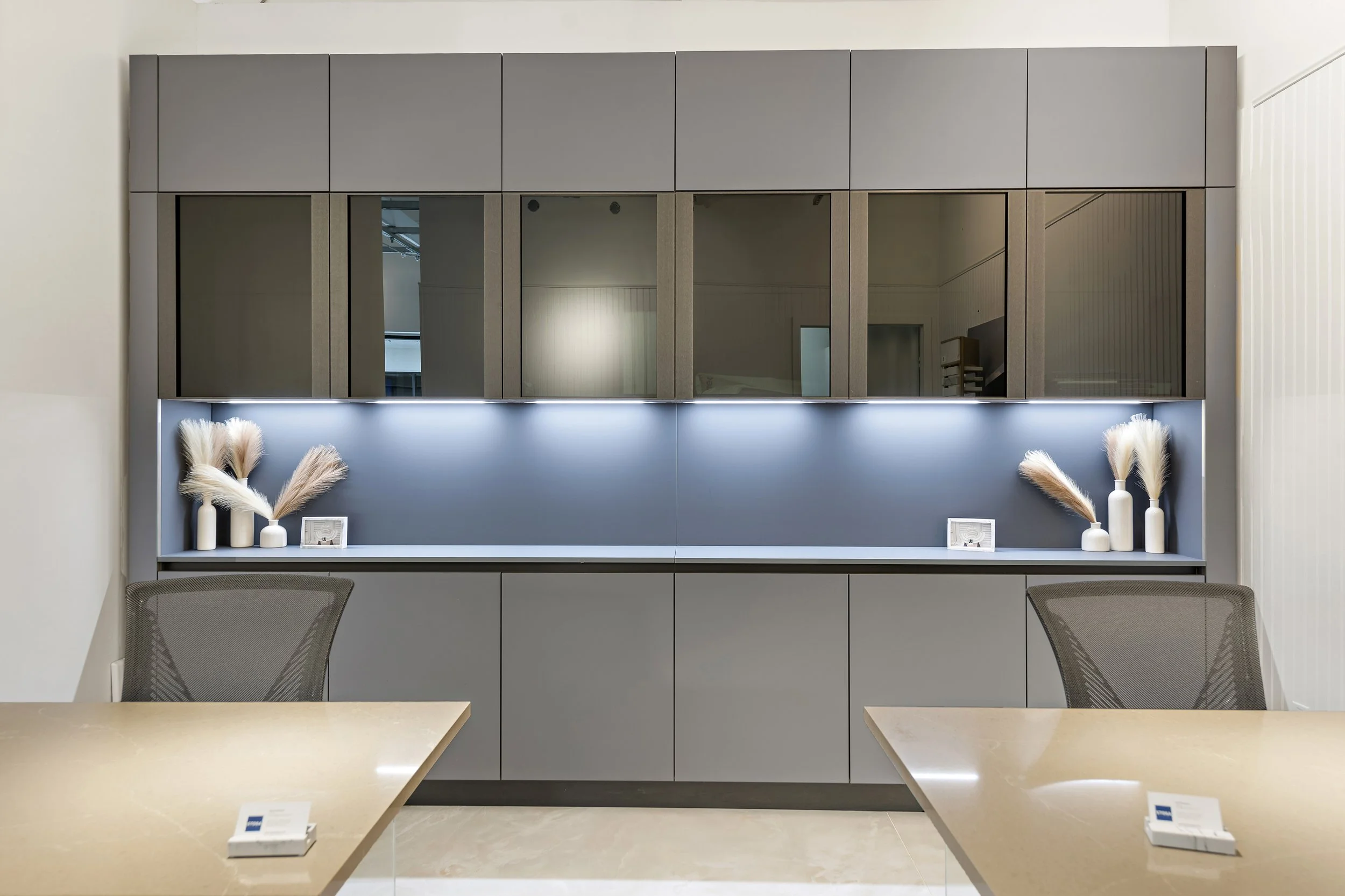 Modern office or conference room with beige tables, gray chairs, and a built-in gray and blue cabinet with decorative vases and framed photos.