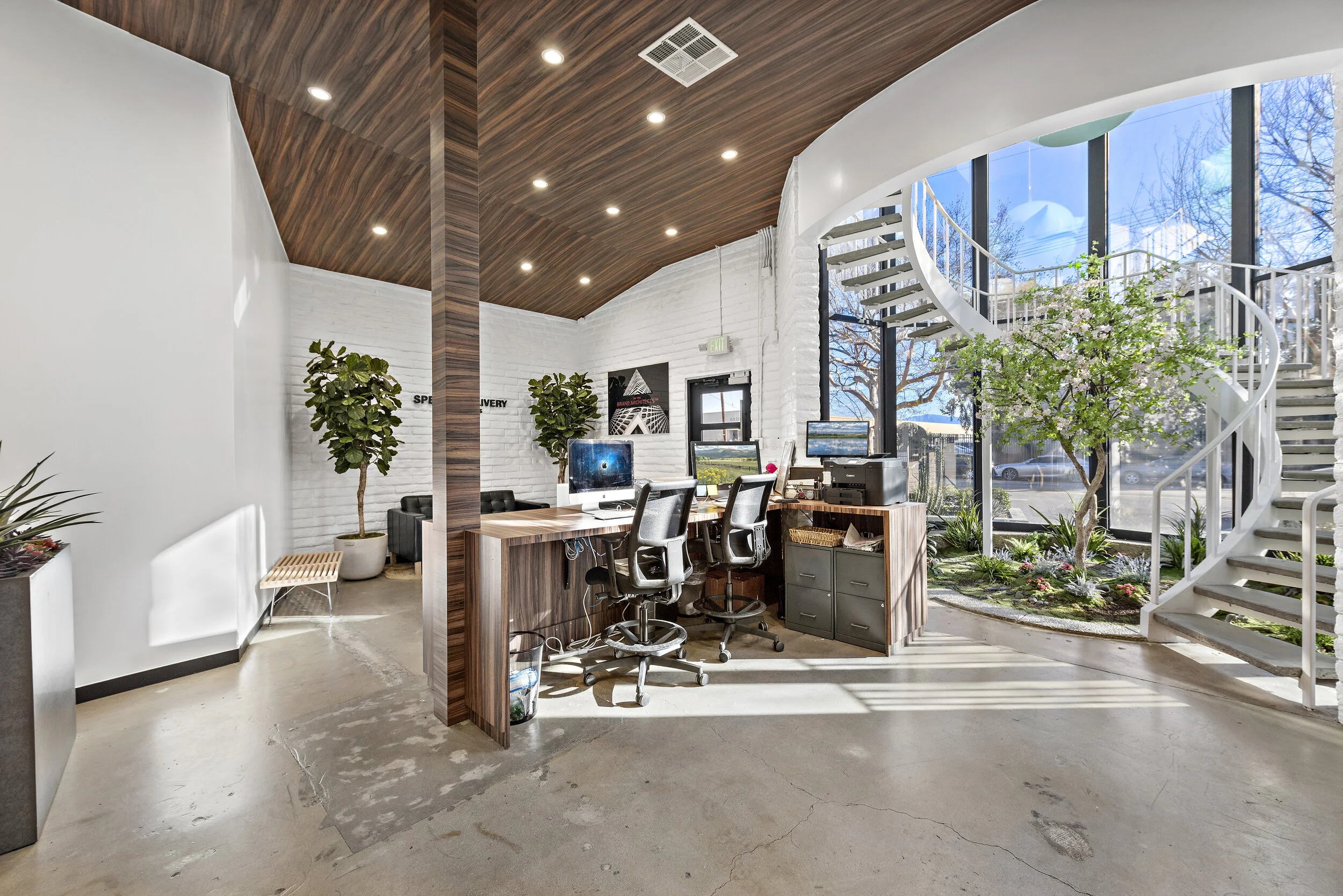 Modern office with a reception desk, computer stations, indoor garden, large window, and a spiral staircase.