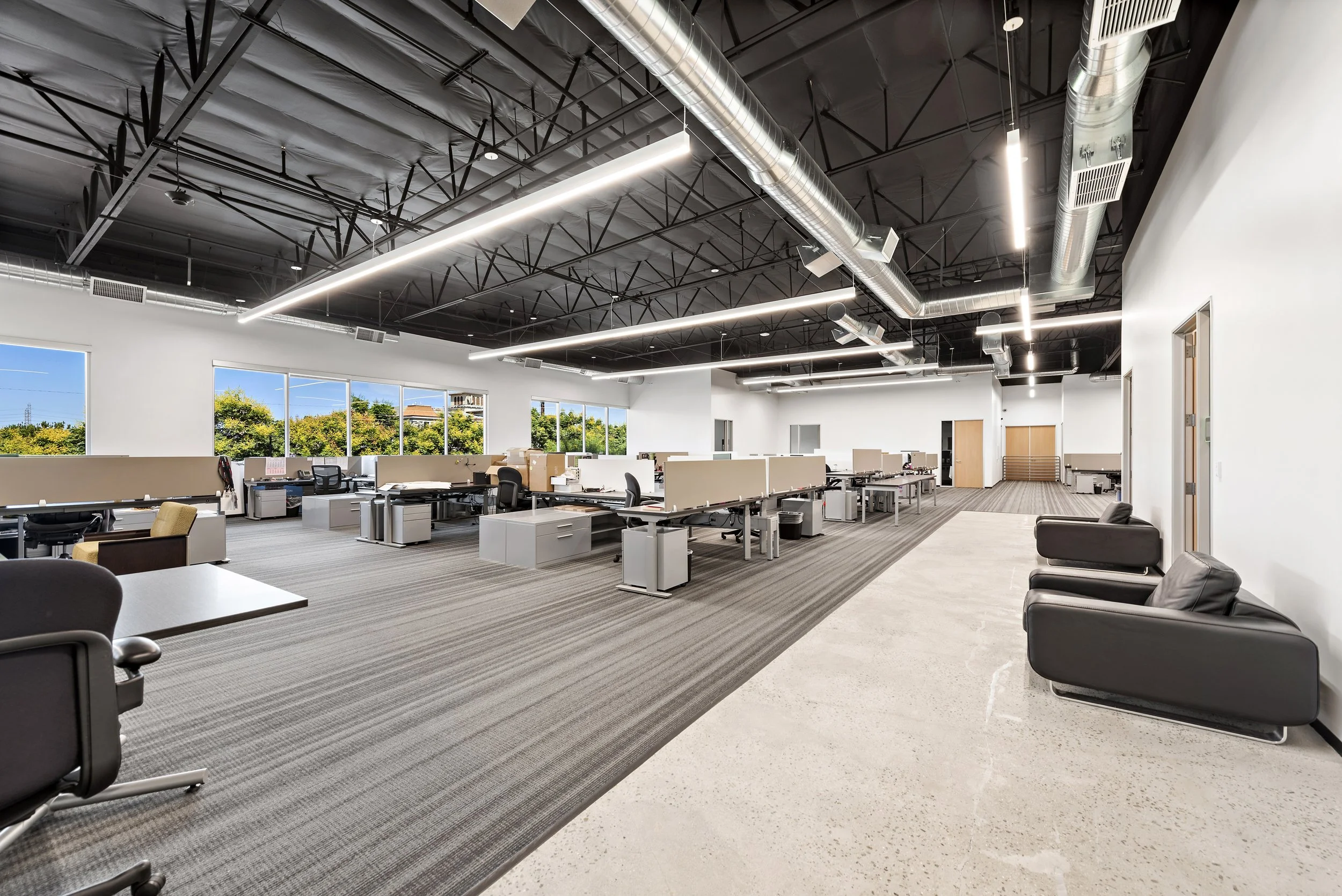 Empty modern office space with rows of desks, some chairs, large windows with trees outside, and high ceiling with exposed ductwork and lighting.