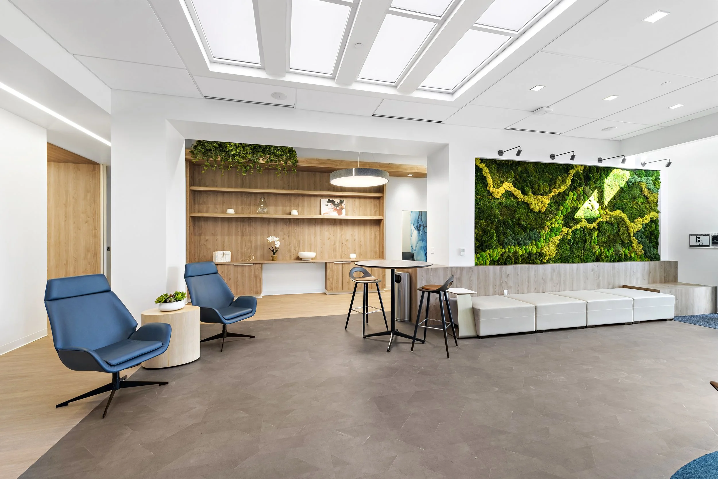 Modern lobby or waiting area with blue lounge chairs, a white cushioned bench, greenery wall art, wooden accents, and ceiling skylights.