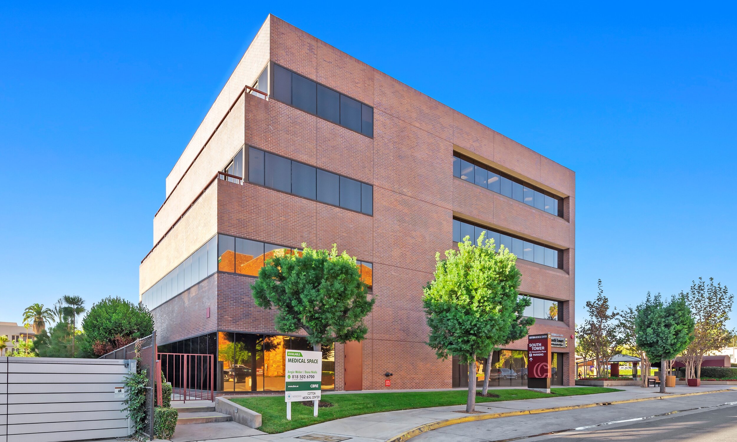 Modern brick office building with large windows, surrounded by trees and a sidewalk, under a blue sky.