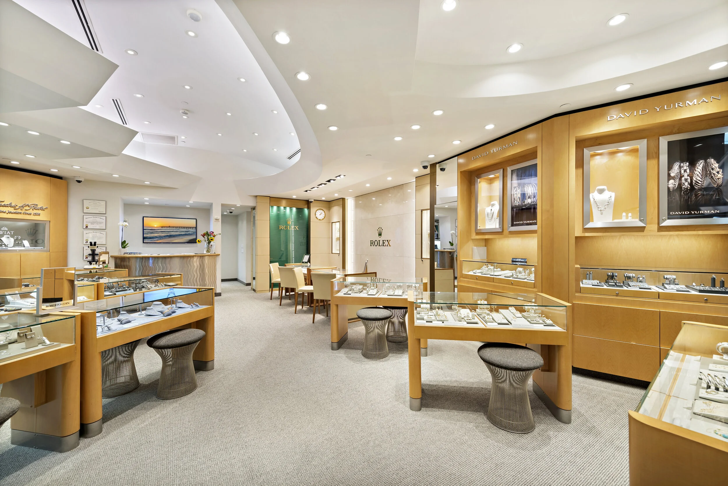 Inside a luxury jewelry store featuring display cases with jewelry, watches, and accessories, branded with Rolex and David Yurman logos, with seating areas and bright lighting.