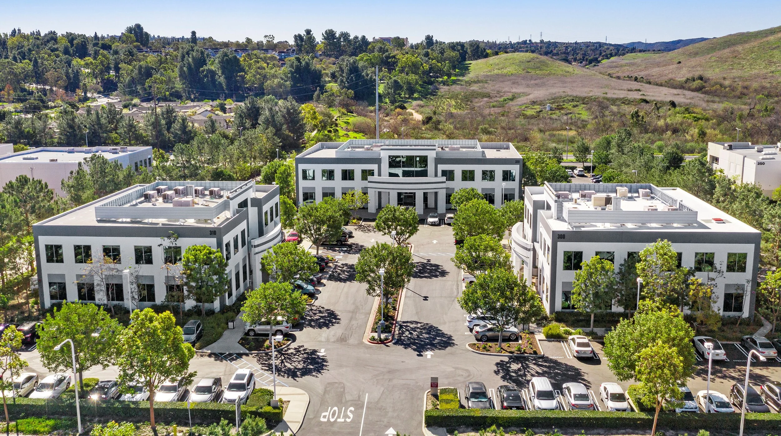 An aerial view of three modern white office buildings with parking lots, surrounded by trees and green landscape, with hills and a forest in the background.