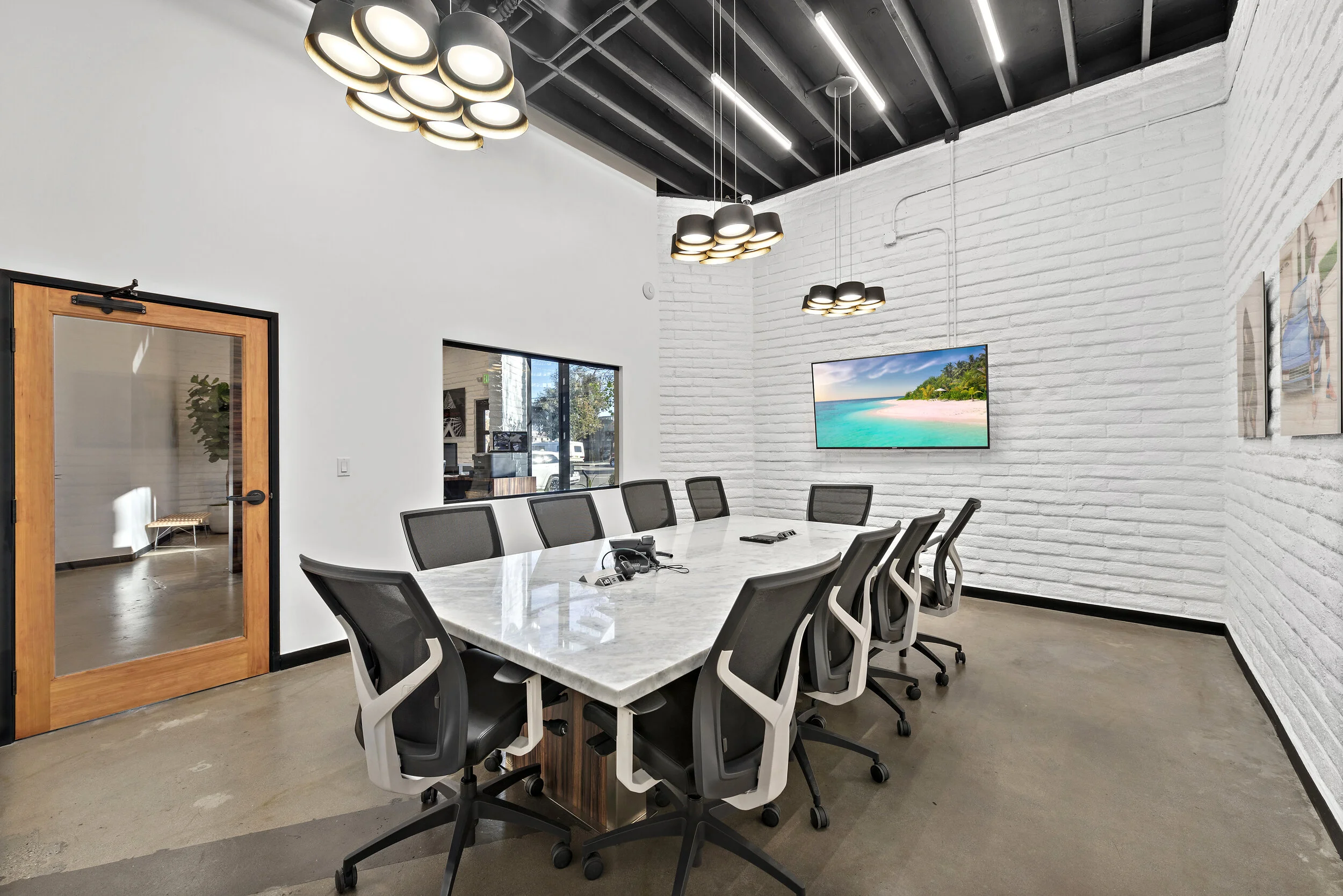 A modern conference room with a large white marble table surrounded by black and white office chairs, a wall-mounted flat-screen TV showing a beach scene, a pendant light fixture overhead, and a glass door leading to another room.