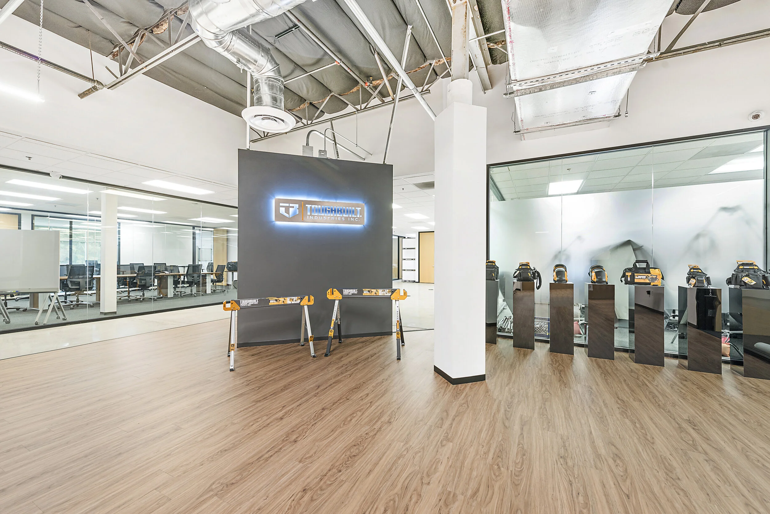 Inside an office with a wall displaying the ToughBuilt Industries Inc. logo illuminated, surrounded by a glass wall with workstations, and a rack of power tools on black pedestals in front of a mirror.