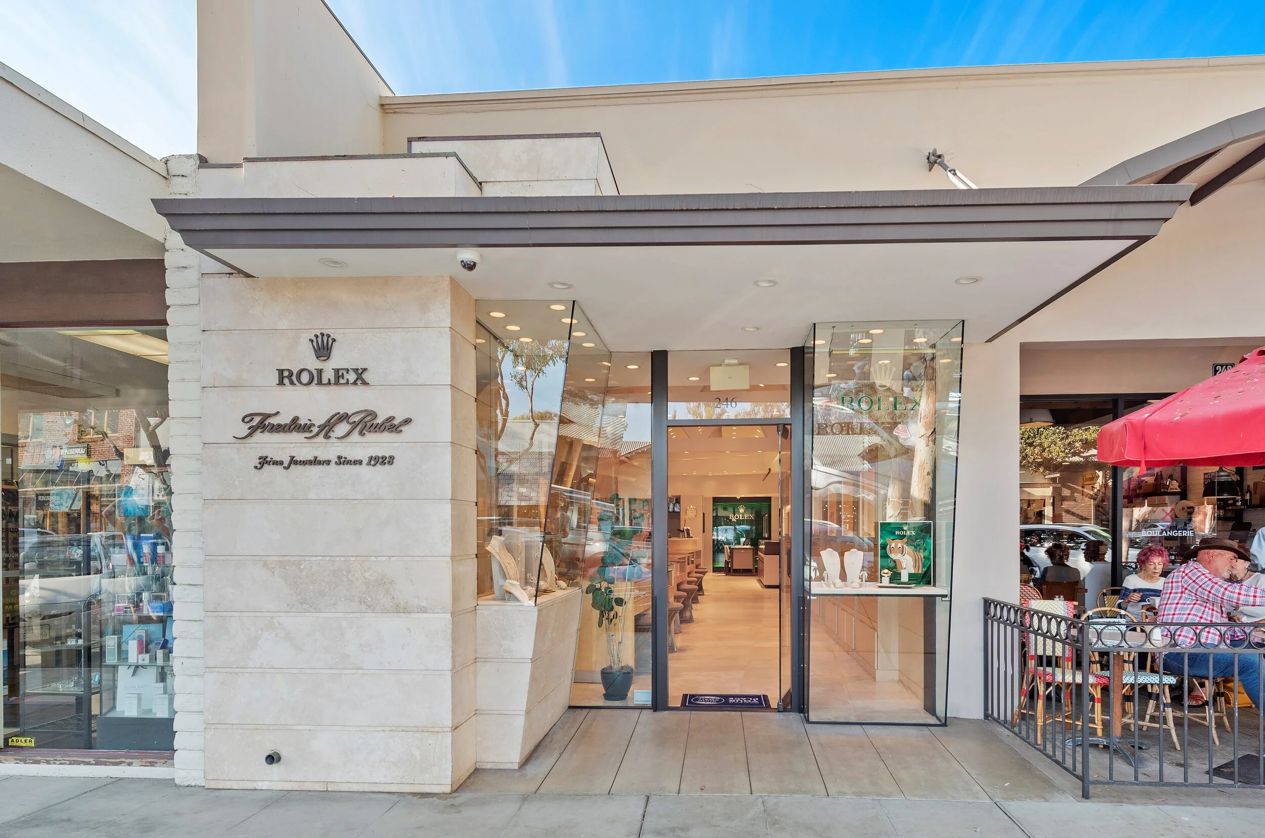 Exterior of a Rolex jewelry store with a white stone facade, glass doors, and display windows, with sidewalk and outdoor seating area to the right.