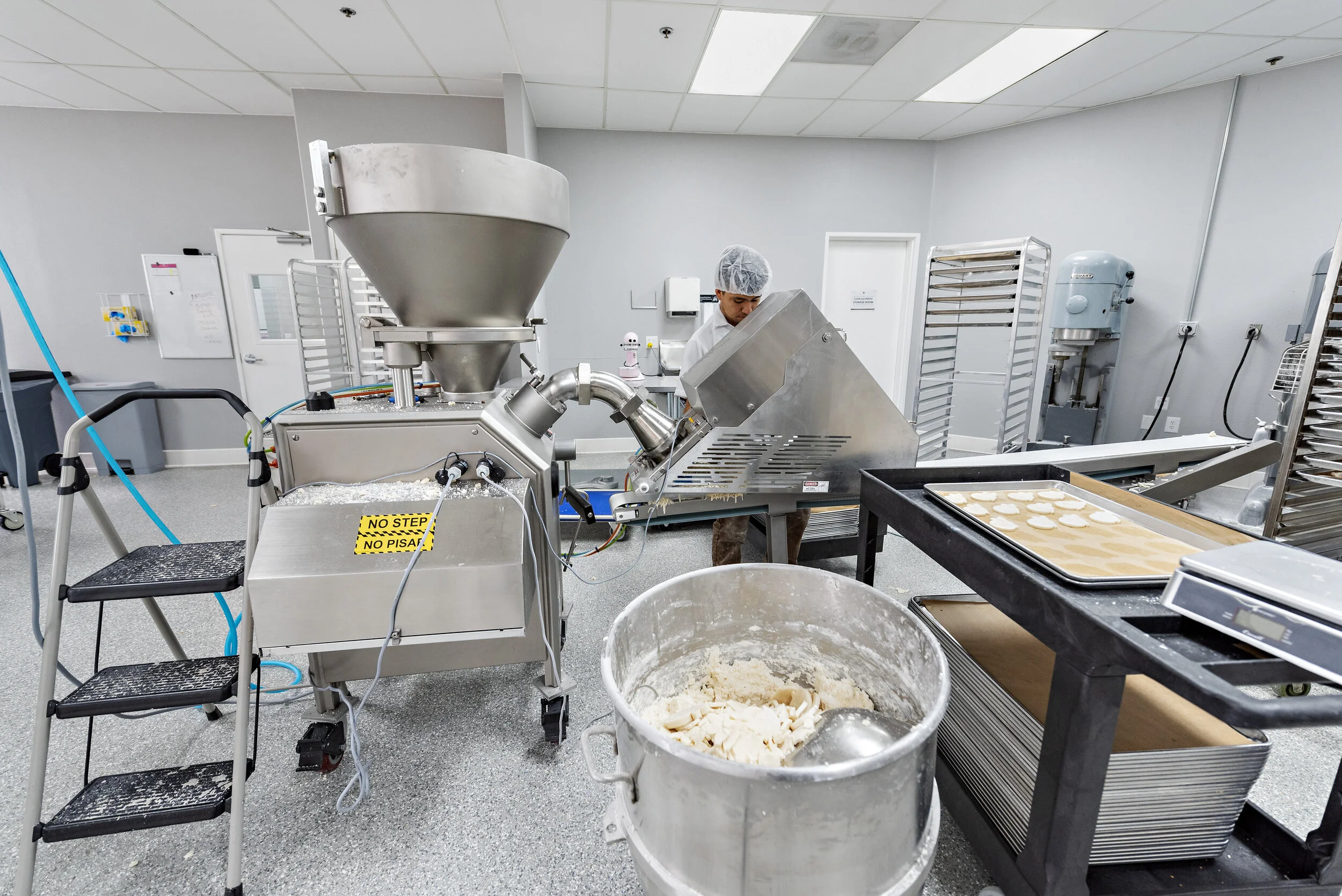 A person working with industrial food processing machinery in a commercial kitchen or bakery, with trays of dough and ingredients nearby.