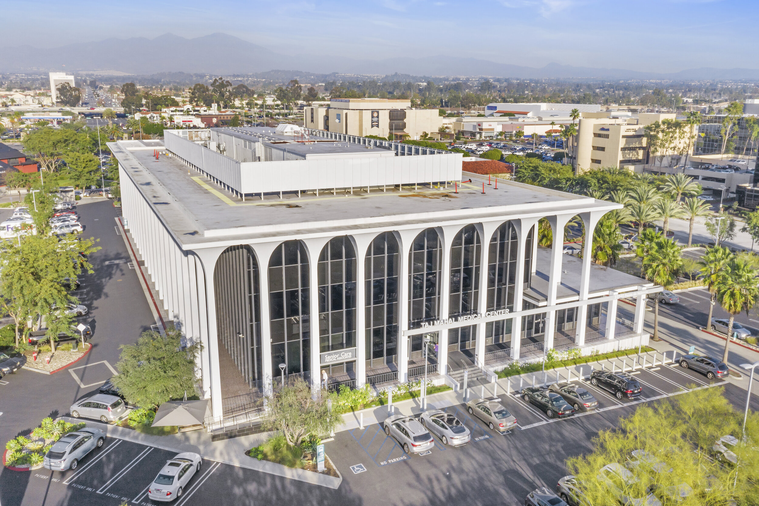 Aerial view of a modern medical center building with tall arched windows, surrounded by a parking lot with cars, palm trees, and other commercial buildings in the background.