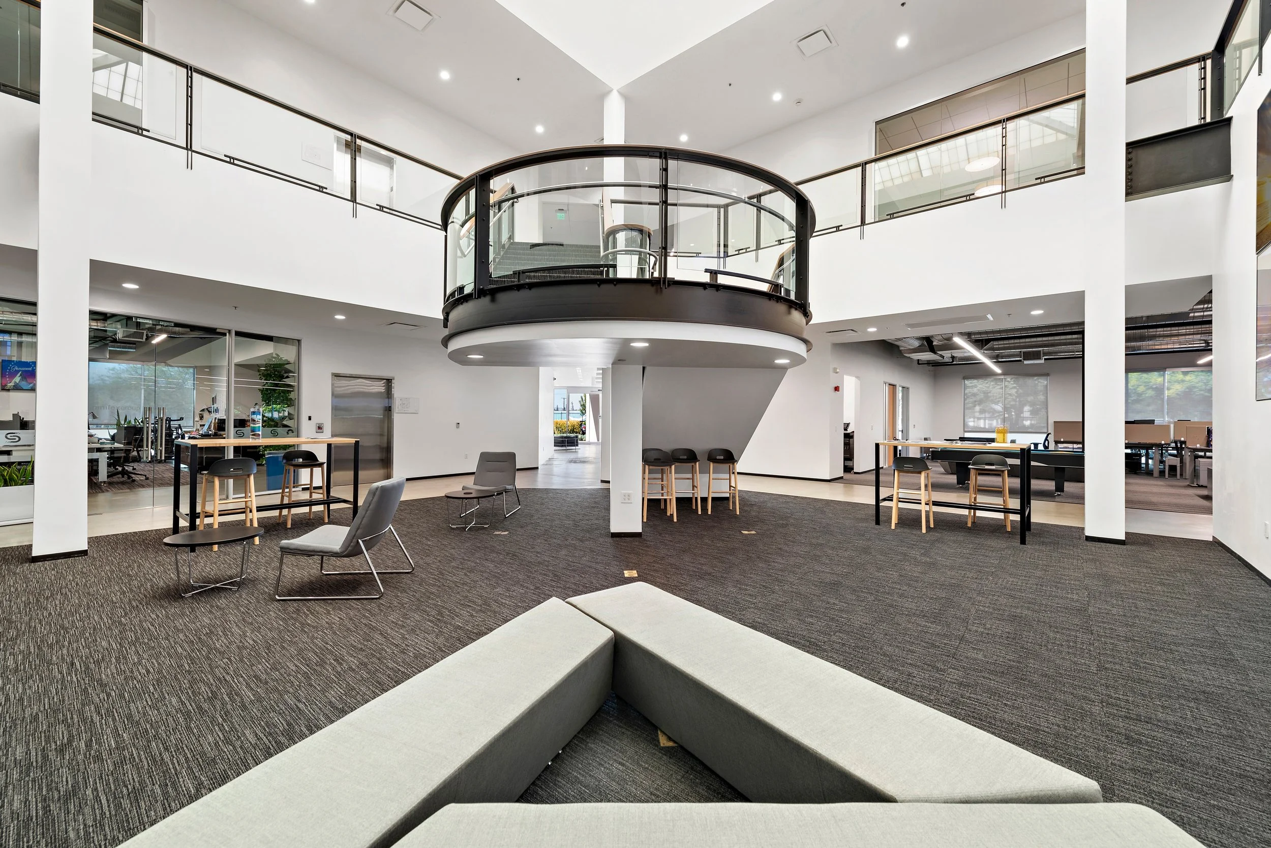 Modern office lobby with seating area, glass railings, and upper balcony, featuring white walls, dark carpet, and ample natural light.