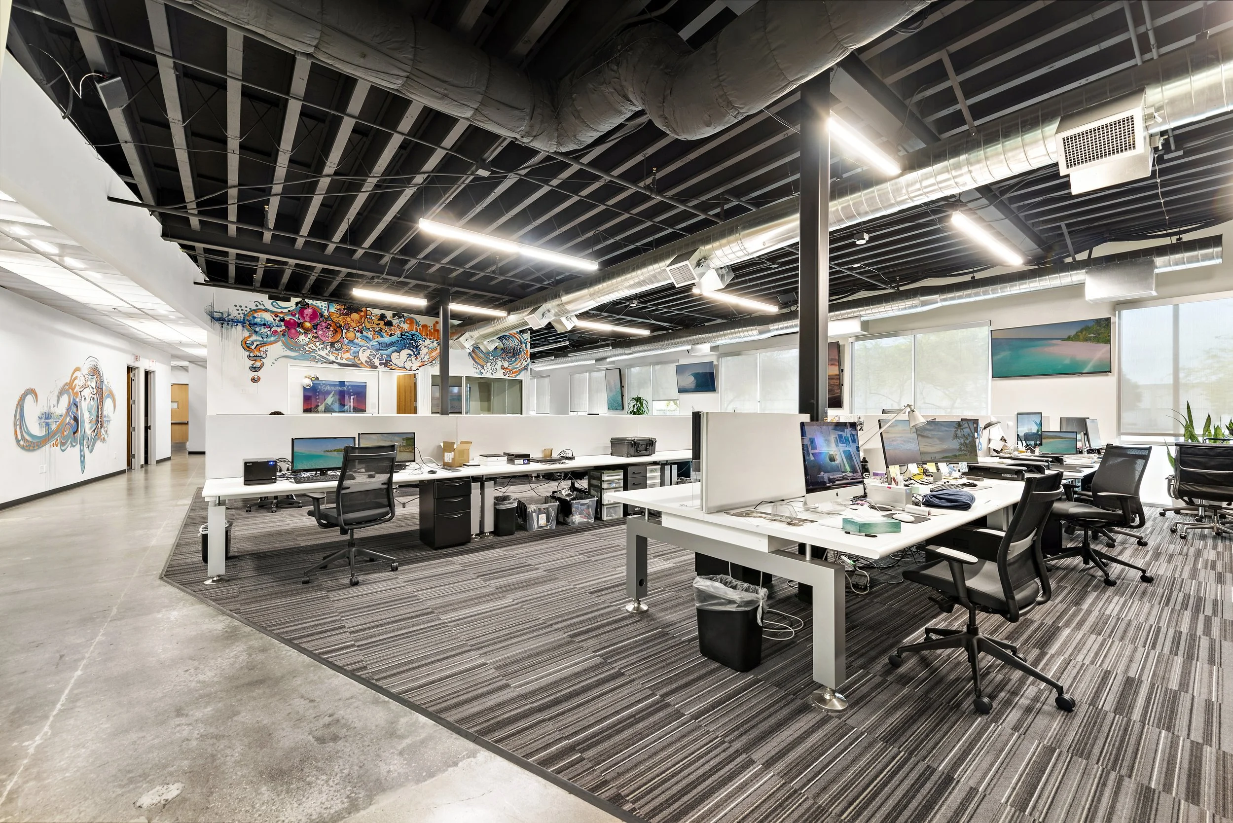 Open office space with multiple desks, black office chairs, computer monitors, and large windows, featuring colorful murals on the walls and an industrial ceiling with exposed ductwork.