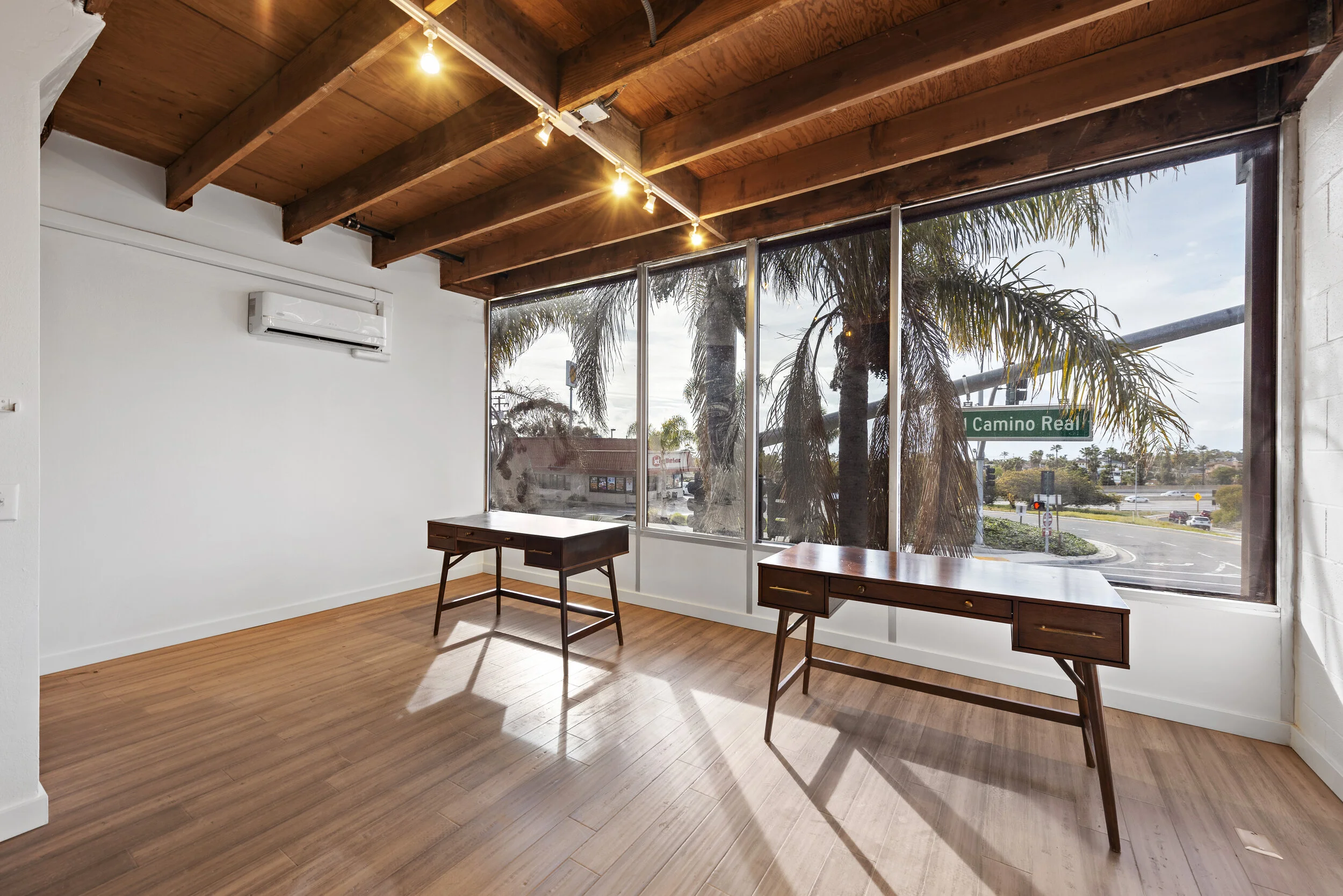 Bright office space with wooden floors, large windows showing palm trees and street view, two wooden desks, and a white wall with an air conditioning unit.