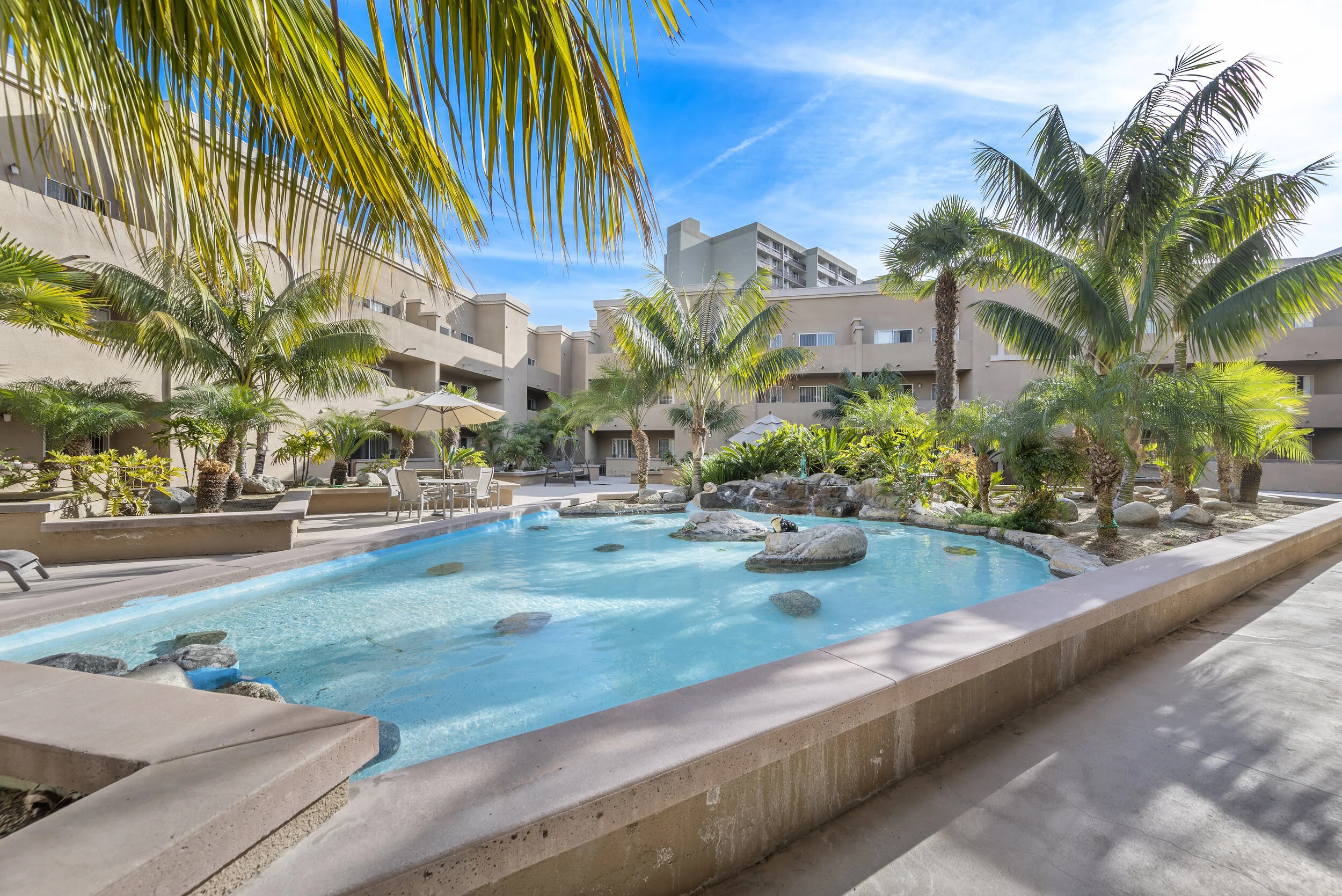 Pool area with palm trees, lounge chairs, and apartment buildings in the background under a bright blue sky.