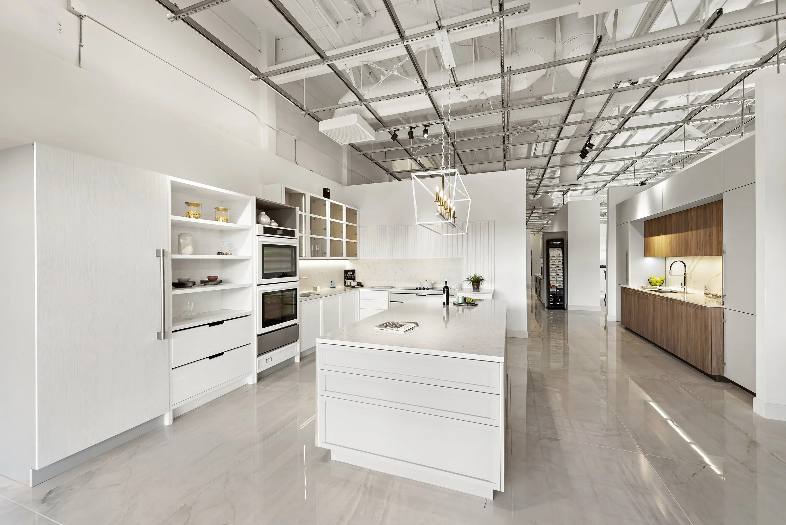 Modern, open-concept kitchen with white cabinets, a marble island, built-in double ovens, and a wood-paneled side cabinet in a spacious, well-lit room with polished floors and an exposed ceiling.