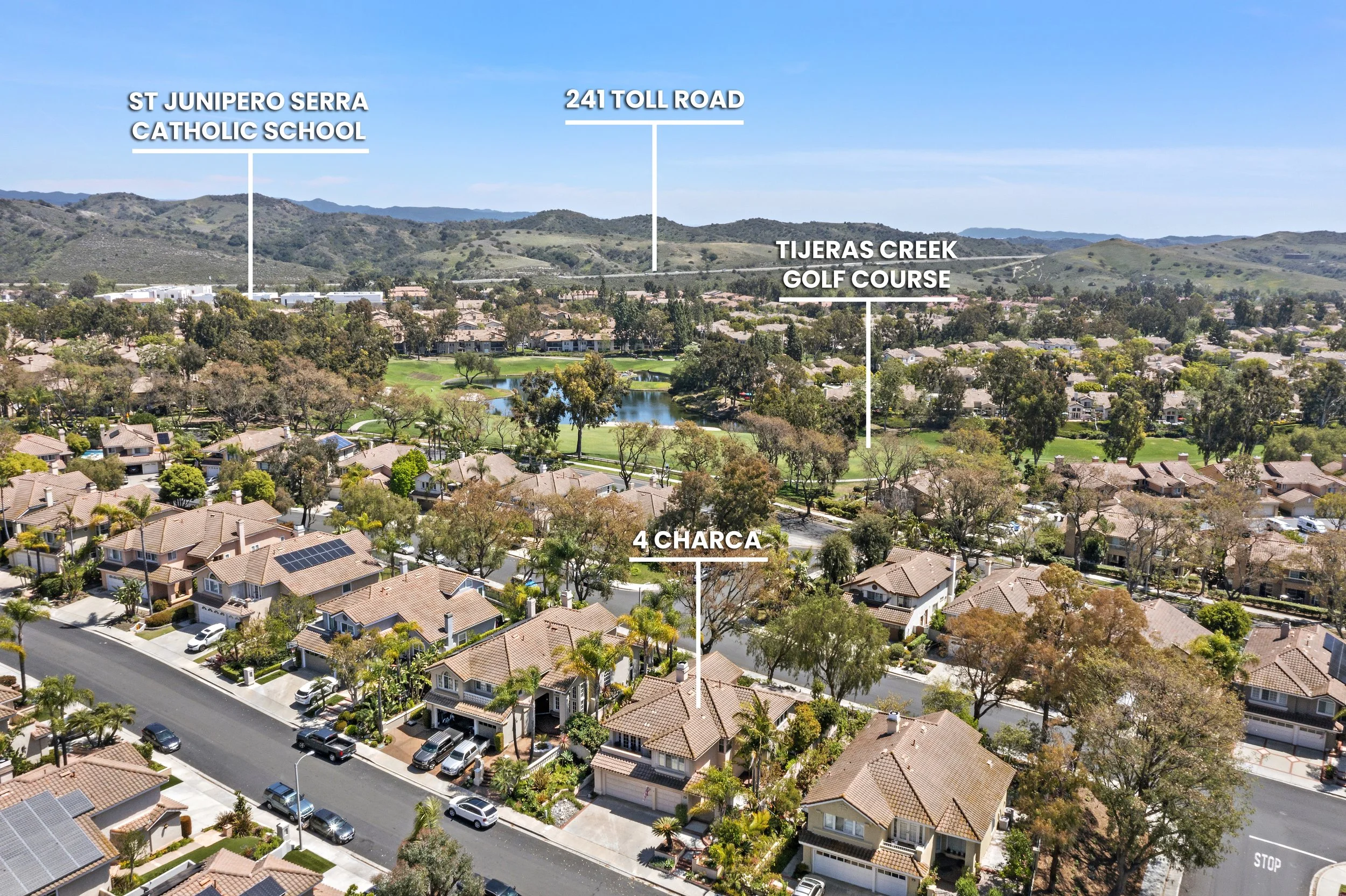 An aerial view of a neighborhood with labeled landmarks: St Junipero Serra Catholic School, 241 Toll Road, Tijeras Creek Golf Course, and 4 Charca, with houses, trees, a pond, and distant hills.