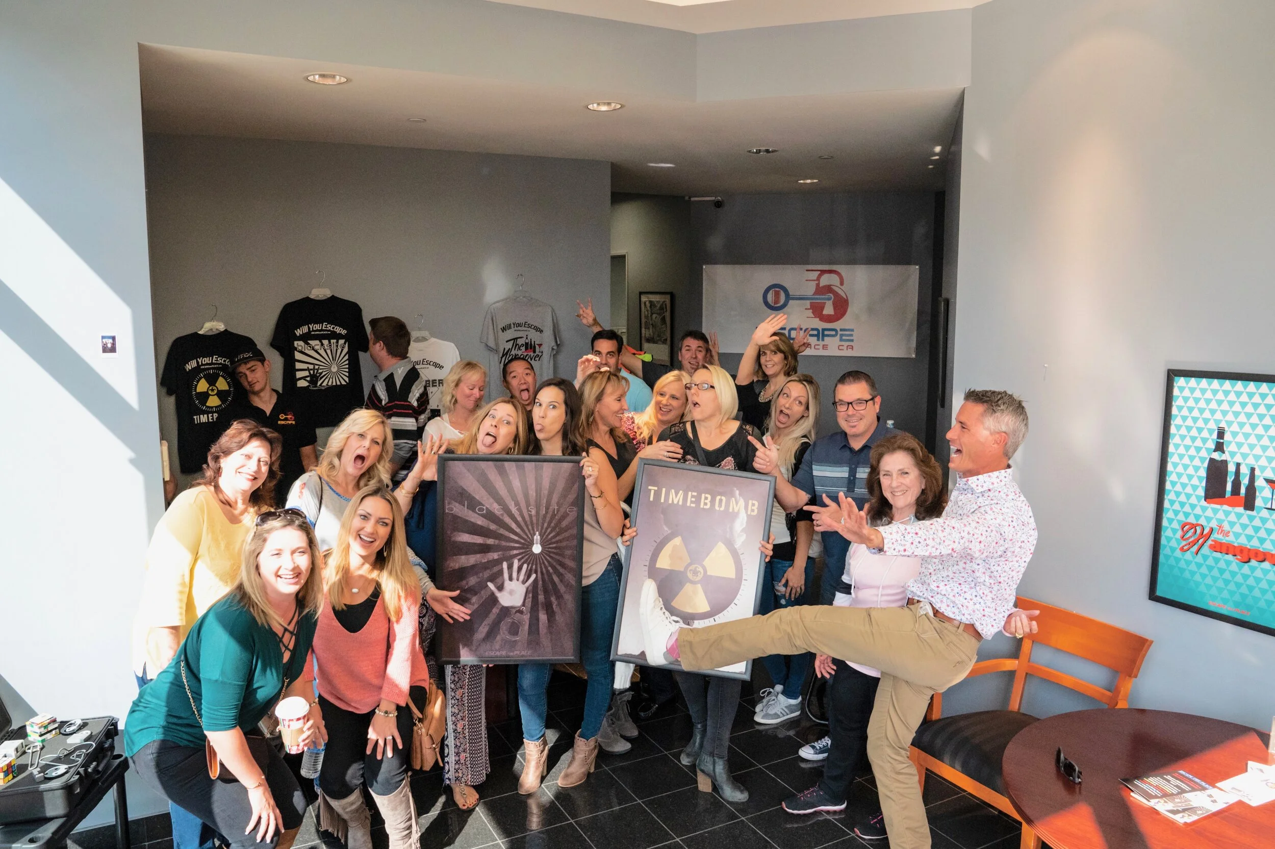 Group of smiling people at a party, holding framed signs labeled 'black list' and 'TIME BOMB', posing enthusiastically, some making gestures, in an indoor space with a wall display and a framed poster.