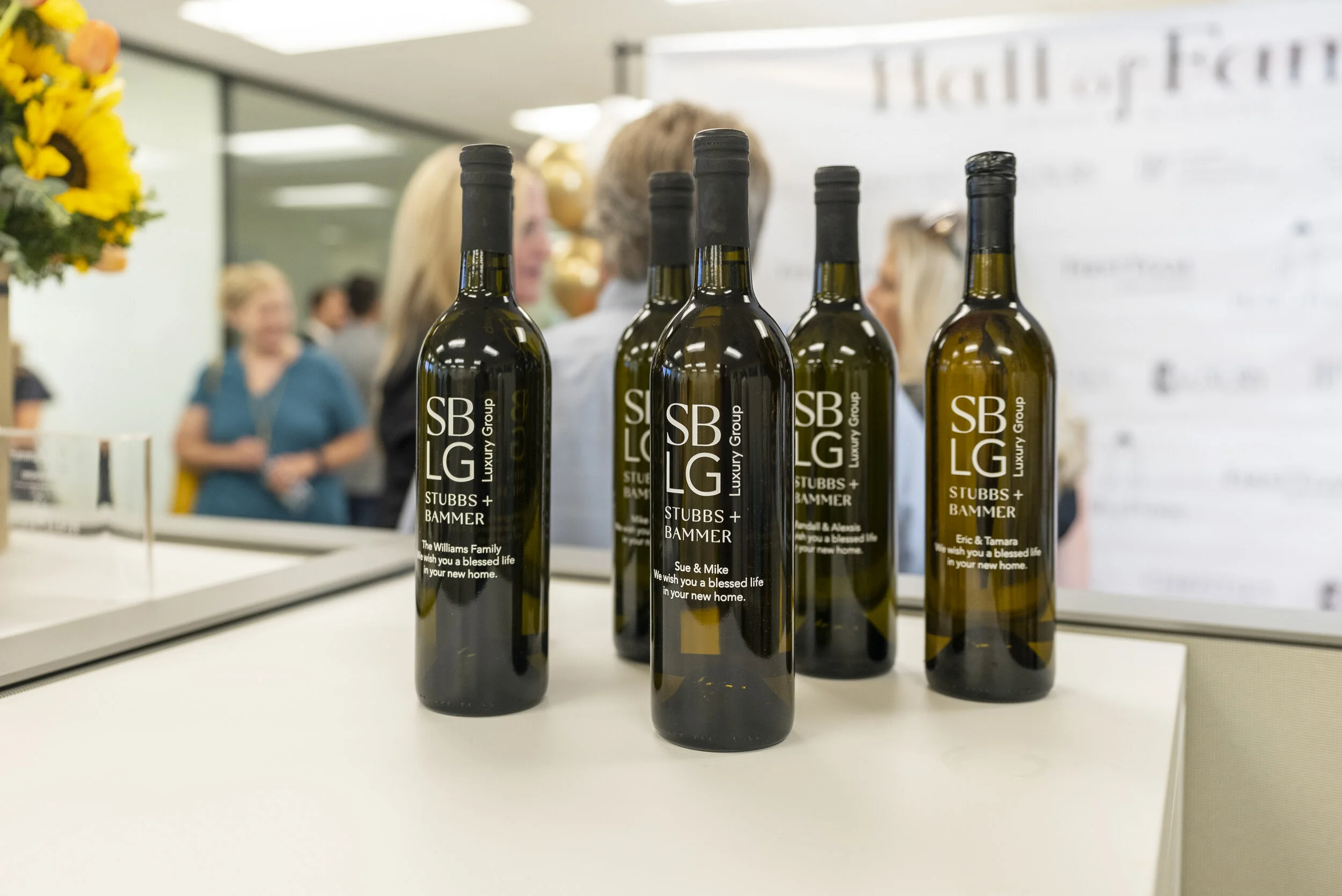 Five dark-colored wine bottles with custom labels are displayed on a white table in an event space. People are blurred in the background.