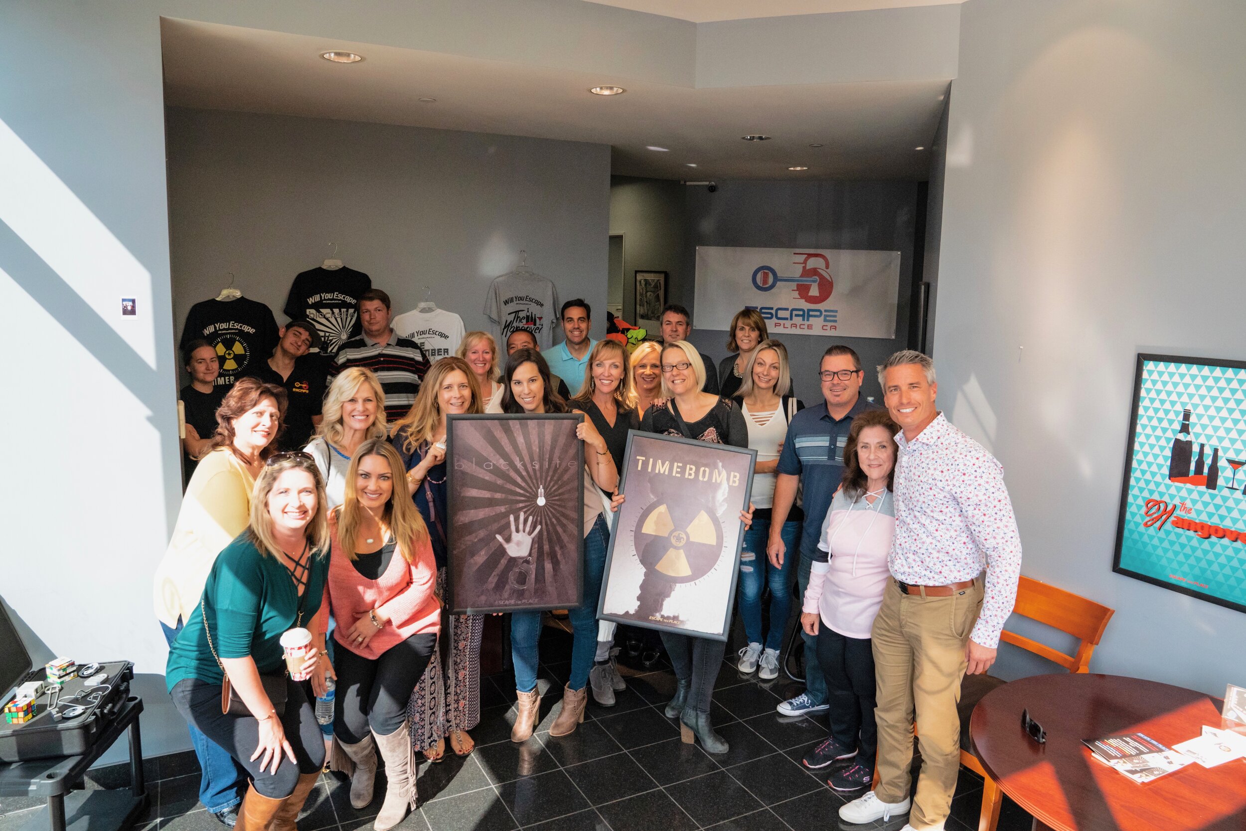 A group of people gathered indoors, smiling and holding framed posters with abstract and radioactive theme designs, in a room with a logo banner on the wall and a poster on a screen, illuminated by overhead lights.
