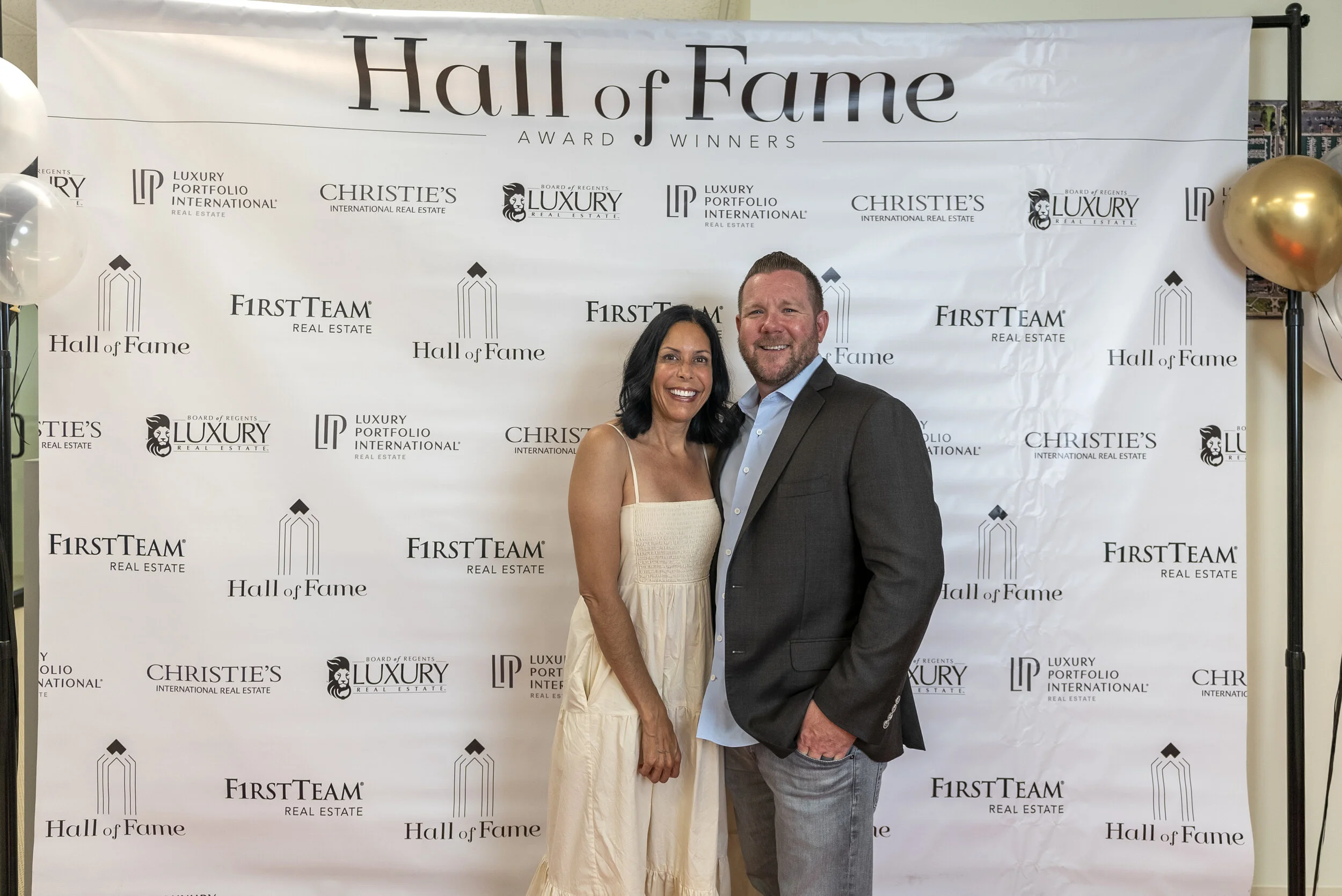 A woman and a man smiling and posing together at a Hall of Fame award event, standing in front of a backdrop with logos and text for Hall of Fame, FirstTeam Real Estate, Luxury Portfolio International, Christie's, and other sponsors.