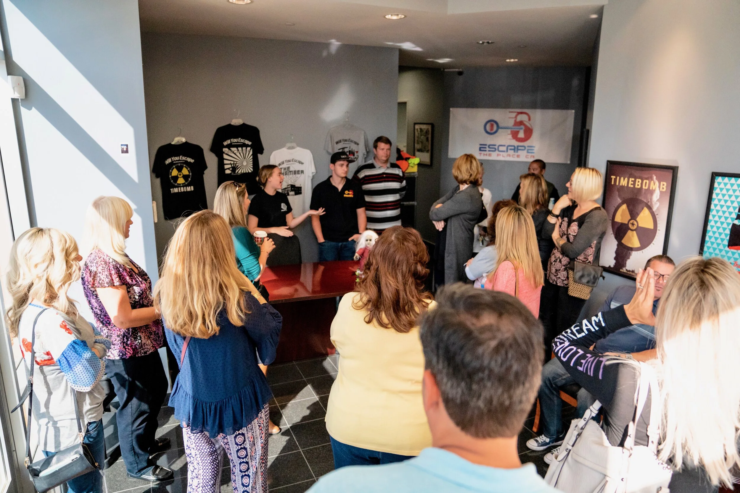 Group of people gathered in a room, listening to a presentation or discussion. T-shirts with radiation symbols and 'Timebomb' graphics are displayed on the wall. Some attendees are standing near the wall, while others are seated or standing around a 
