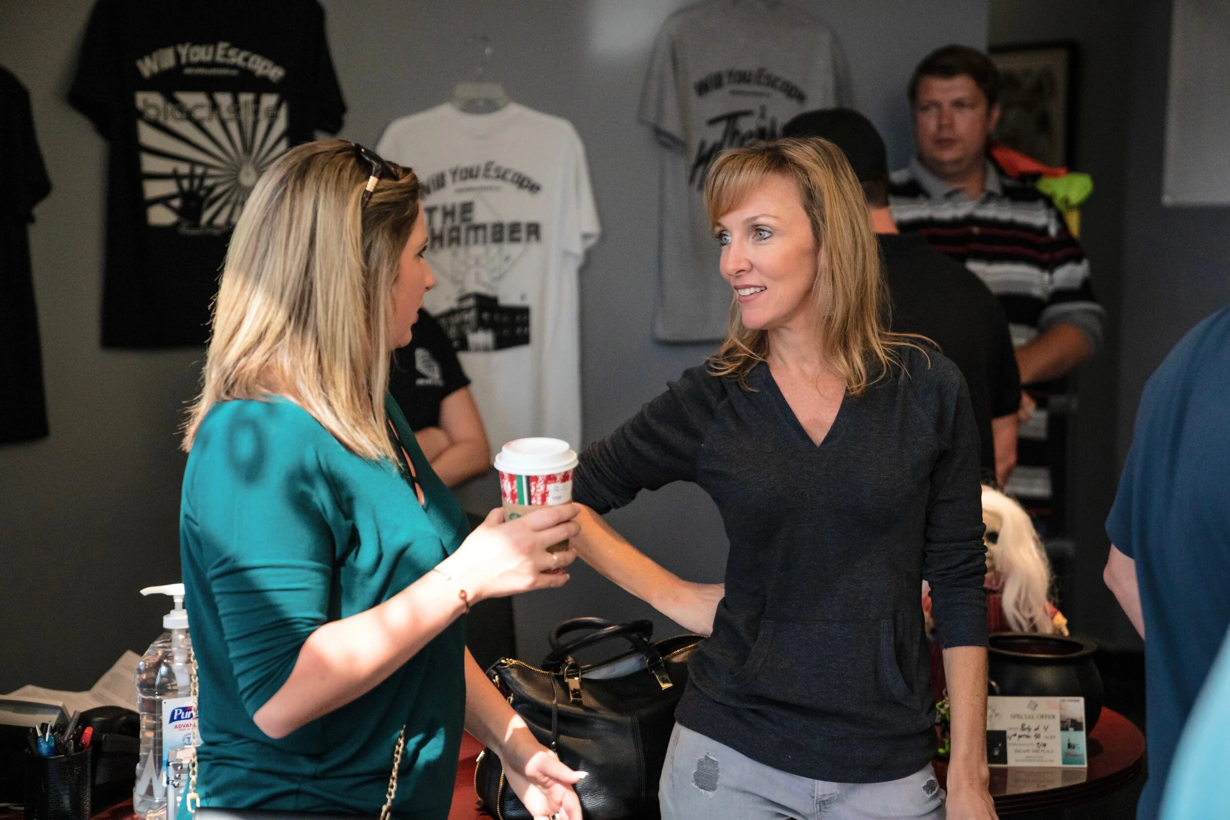Two women conversing at a reception desk, one holding a coffee cup, in a room decorated with t-shirts on the wall.