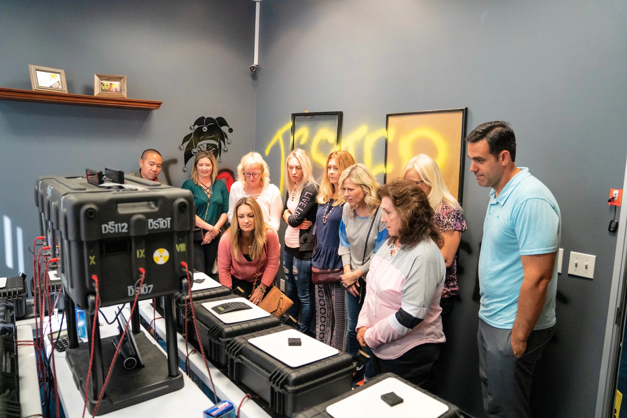 Group of people in a room observing equipment with black cases, on a table, with dark gray walls and neon yellow graffiti-style background and artwork.