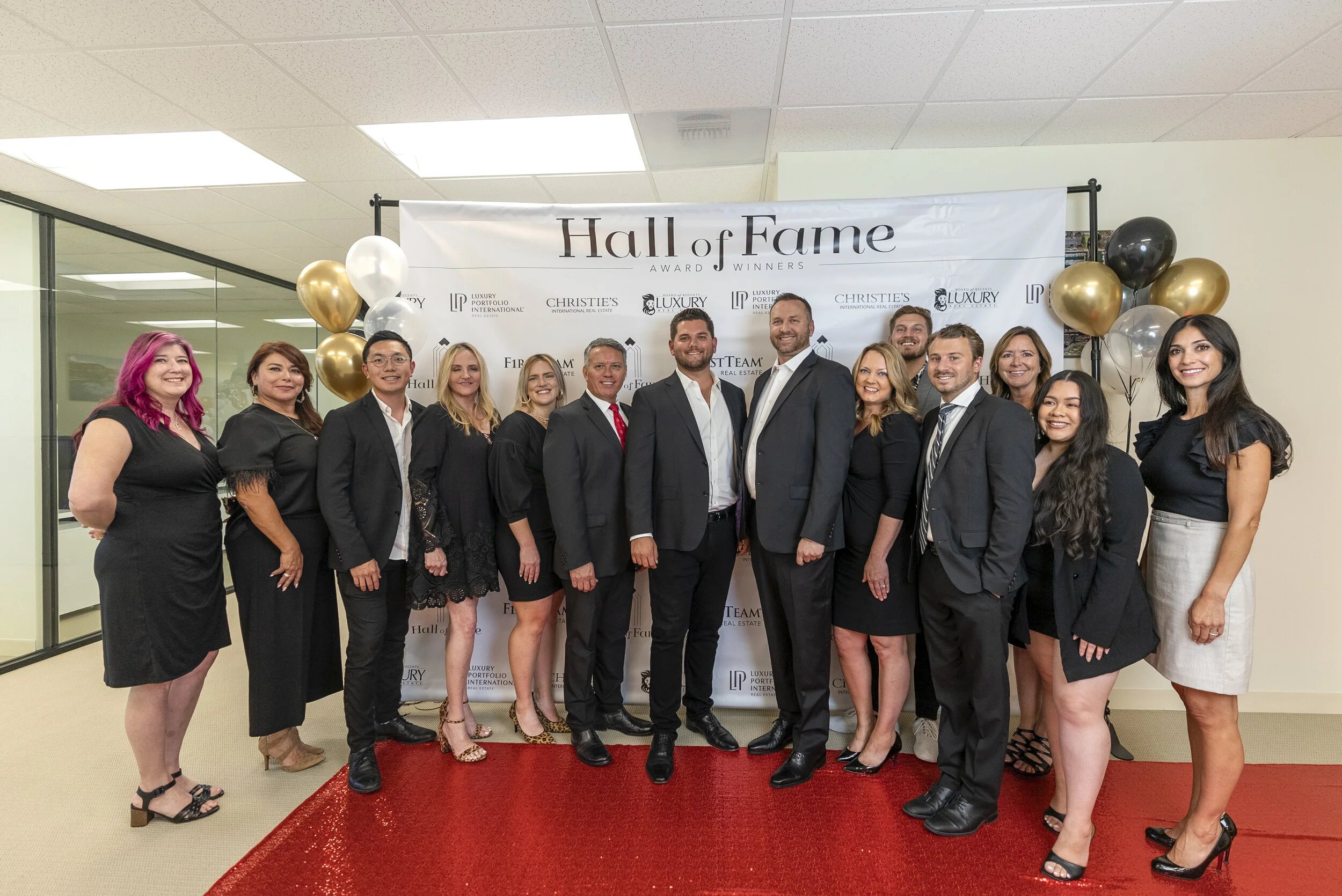 Group of people dressed in formal attire standing on a red carpet at an awards event, with a backdrop reading 'Hall of Fame' and balloons in the background.