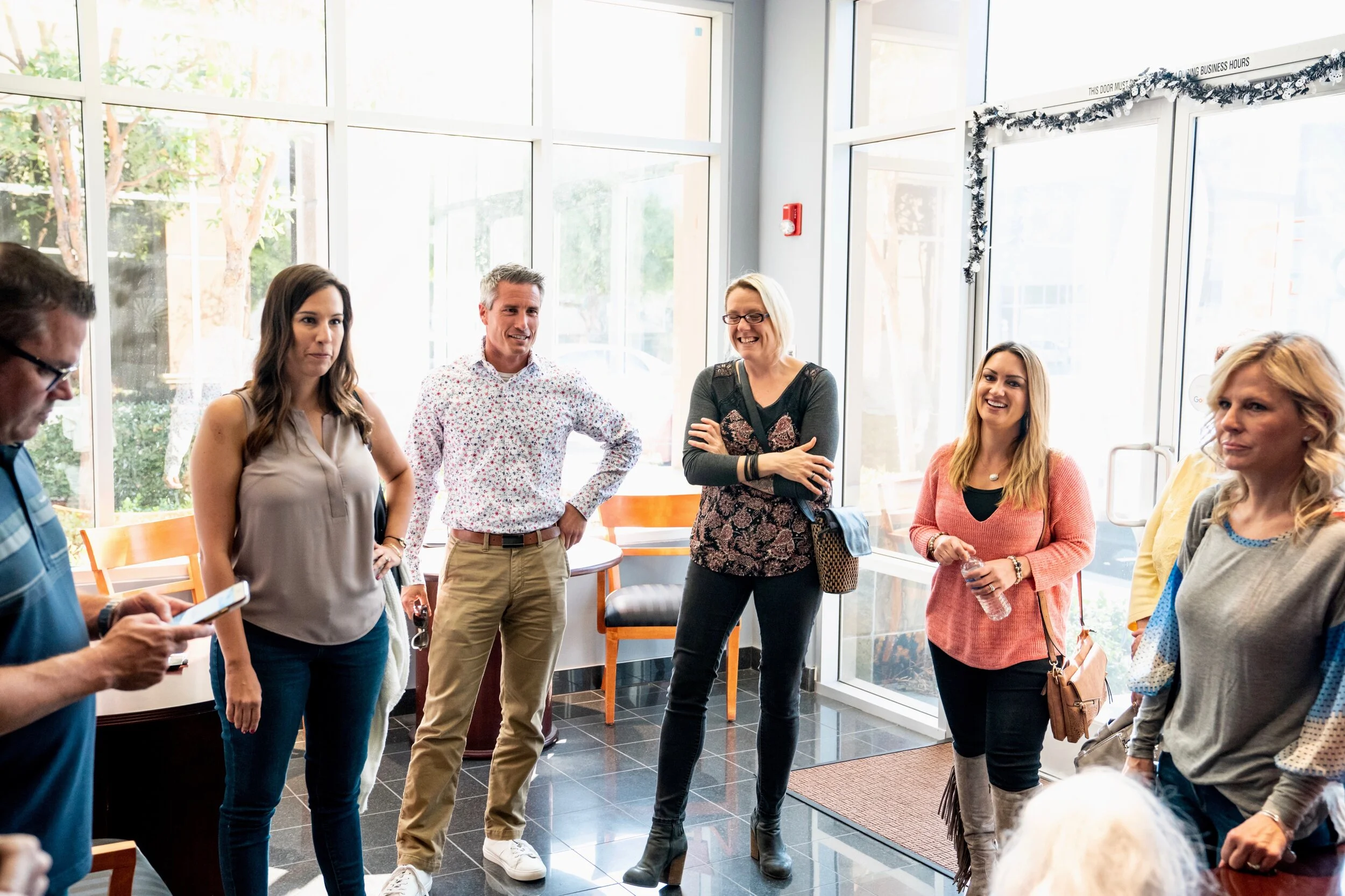 Group of people standing and talking in a brightly lit room with large windows, some smiling and engaged in conversation.