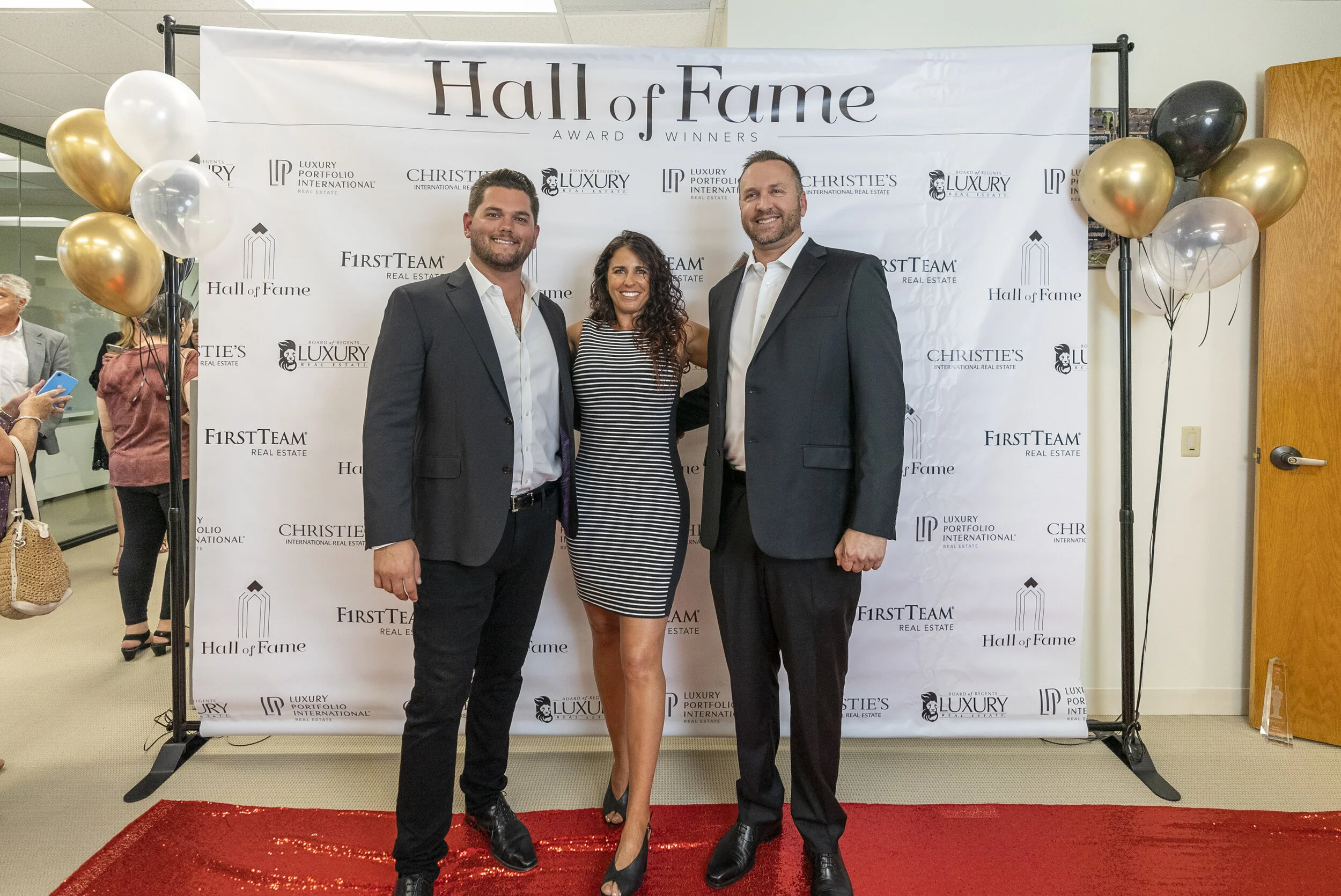 Three people in formal attire standing on a red carpet in front of a backdrop with the words 'Hall of Fame' and logos for real estate companies, surrounded by balloons.