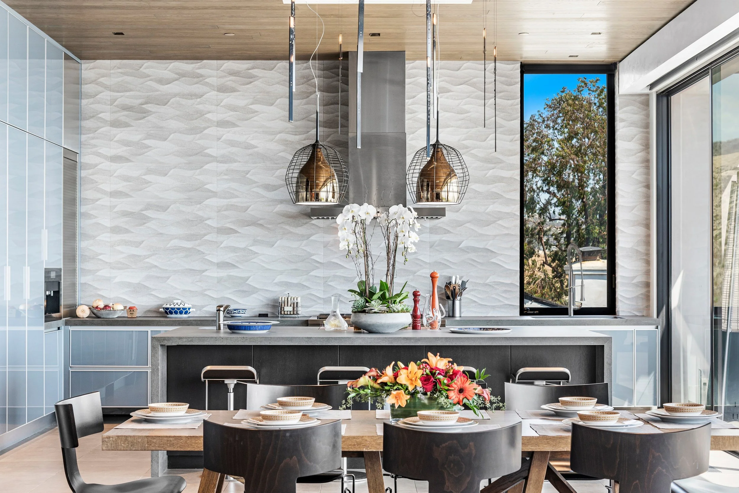 Modern kitchen with a dining table set for six, decorated with a colorful floral centerpiece. The background features a white textured wall, a large window with a view of trees, and a kitchen island with a potted plant, cookware, and utensils.