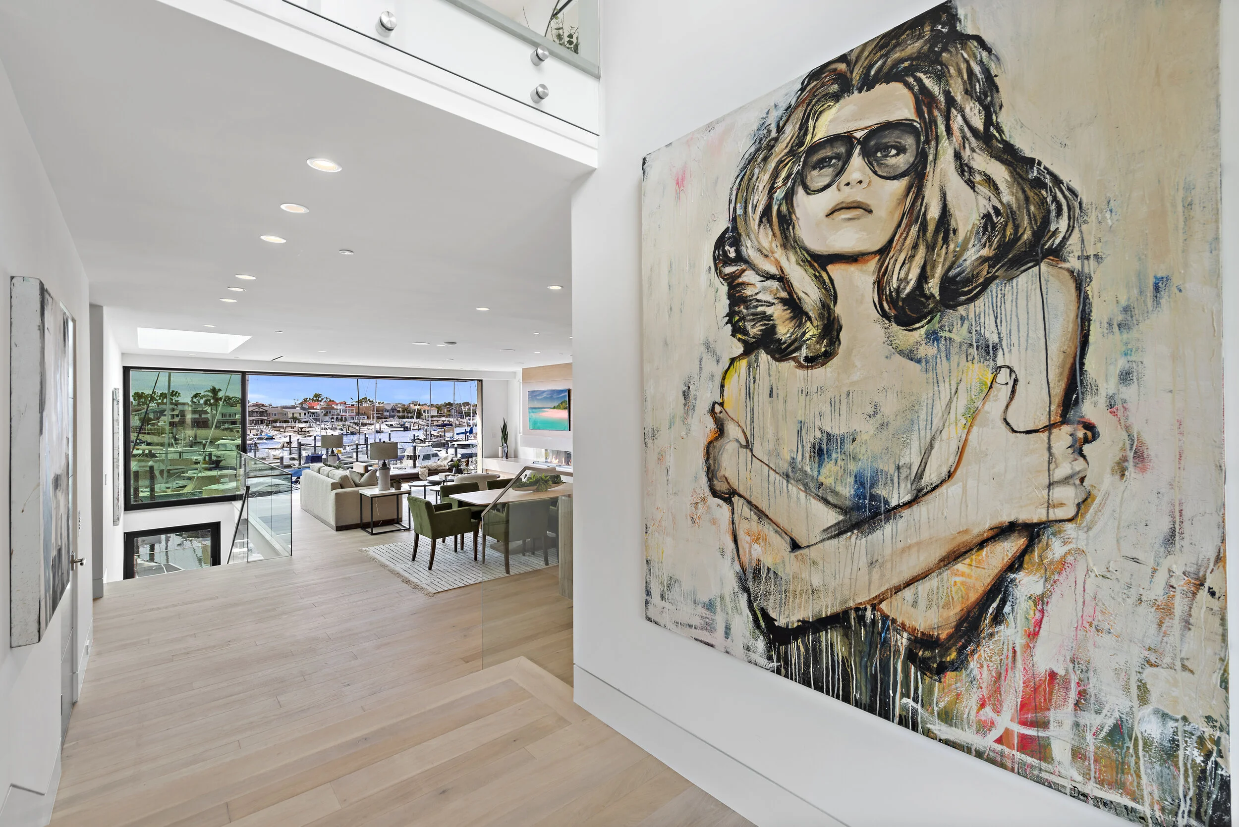 Interior of a modern home featuring a large colorful portrait of a woman with sunglasses, overlooking a living room with sofas, chairs, and a view of a marina with boats through large windows.