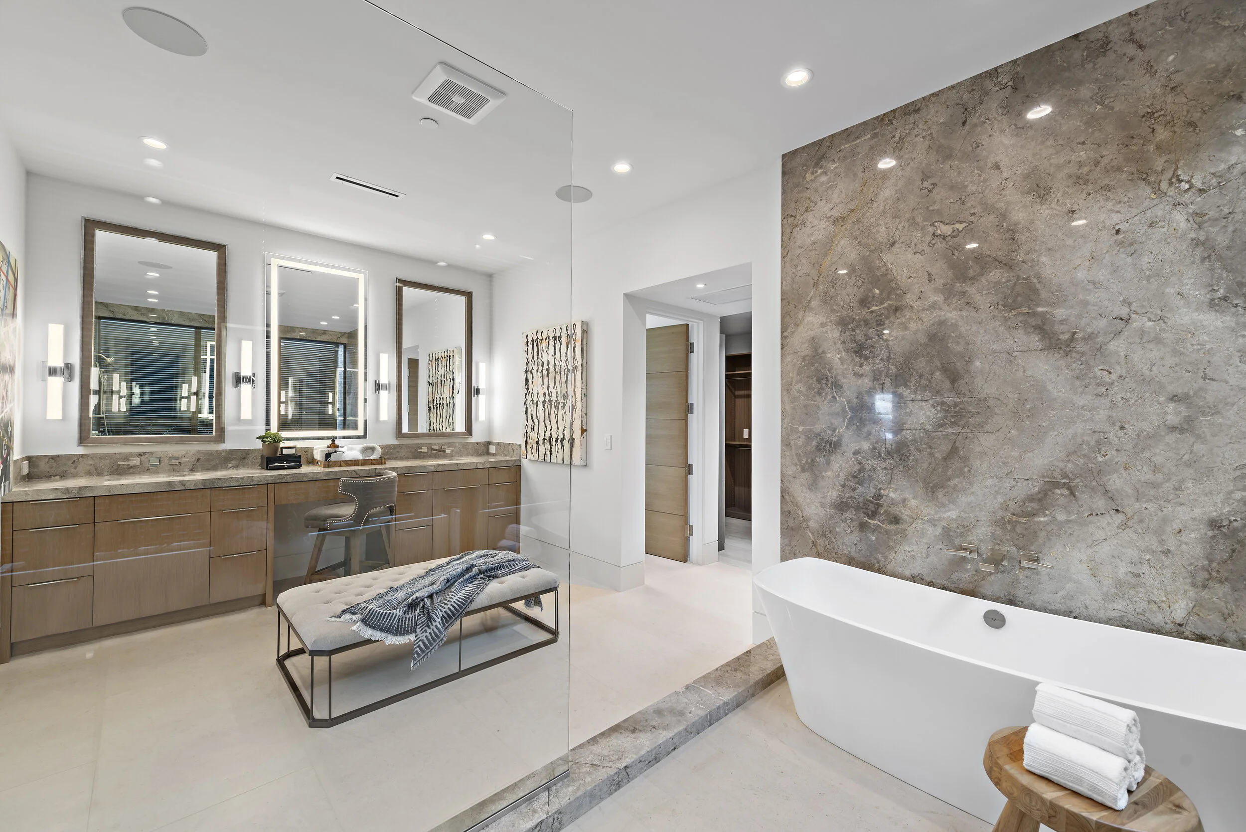 Modern bathroom with a large freestanding bathtub, marble accent wall, vanity with three mirrors, and a wooden bench with folded towels.