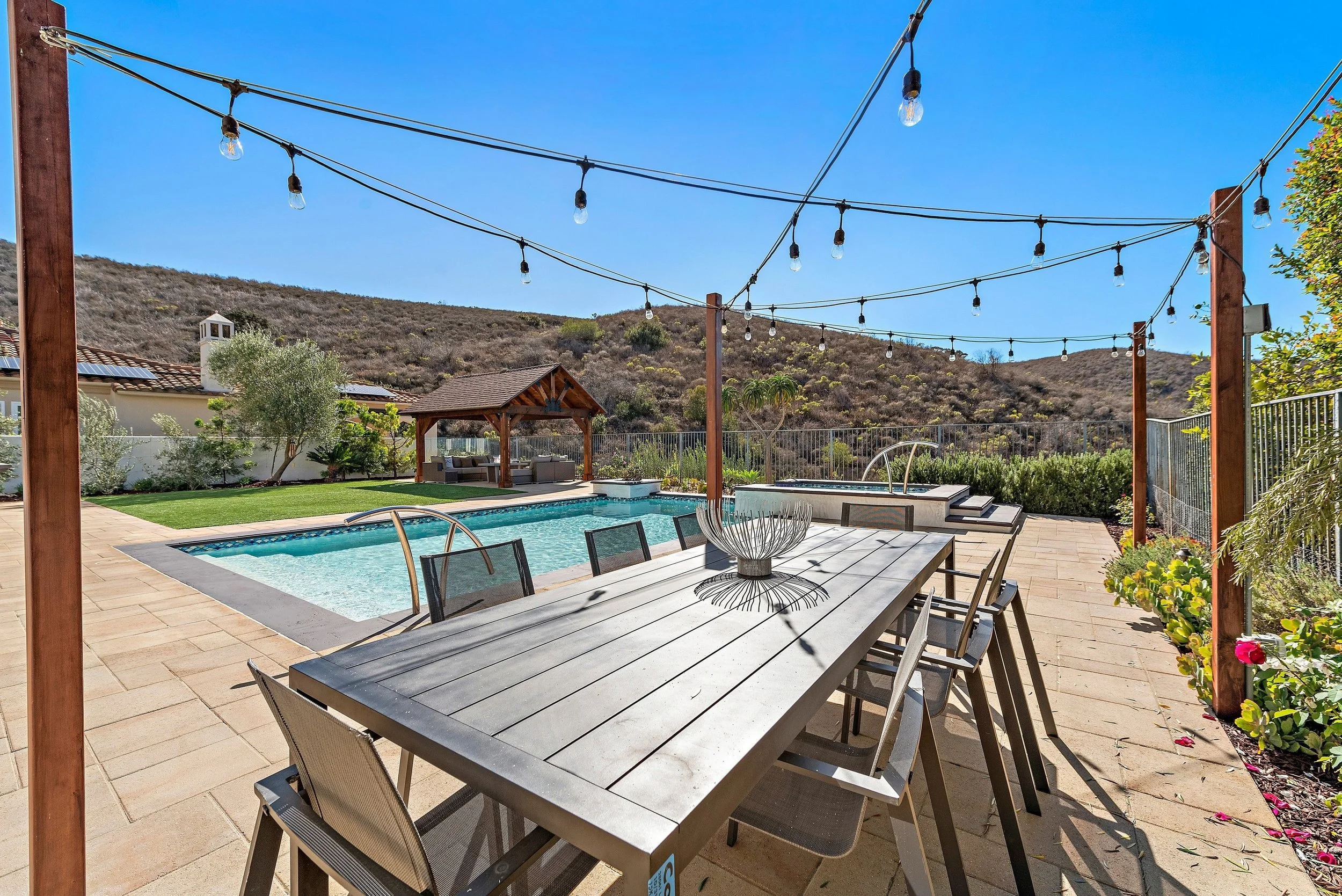 Outdoor backyard with swimming pool, wooden patio table with chairs, string lights, and a gazebo in the background, surrounded by hills and clear blue sky.