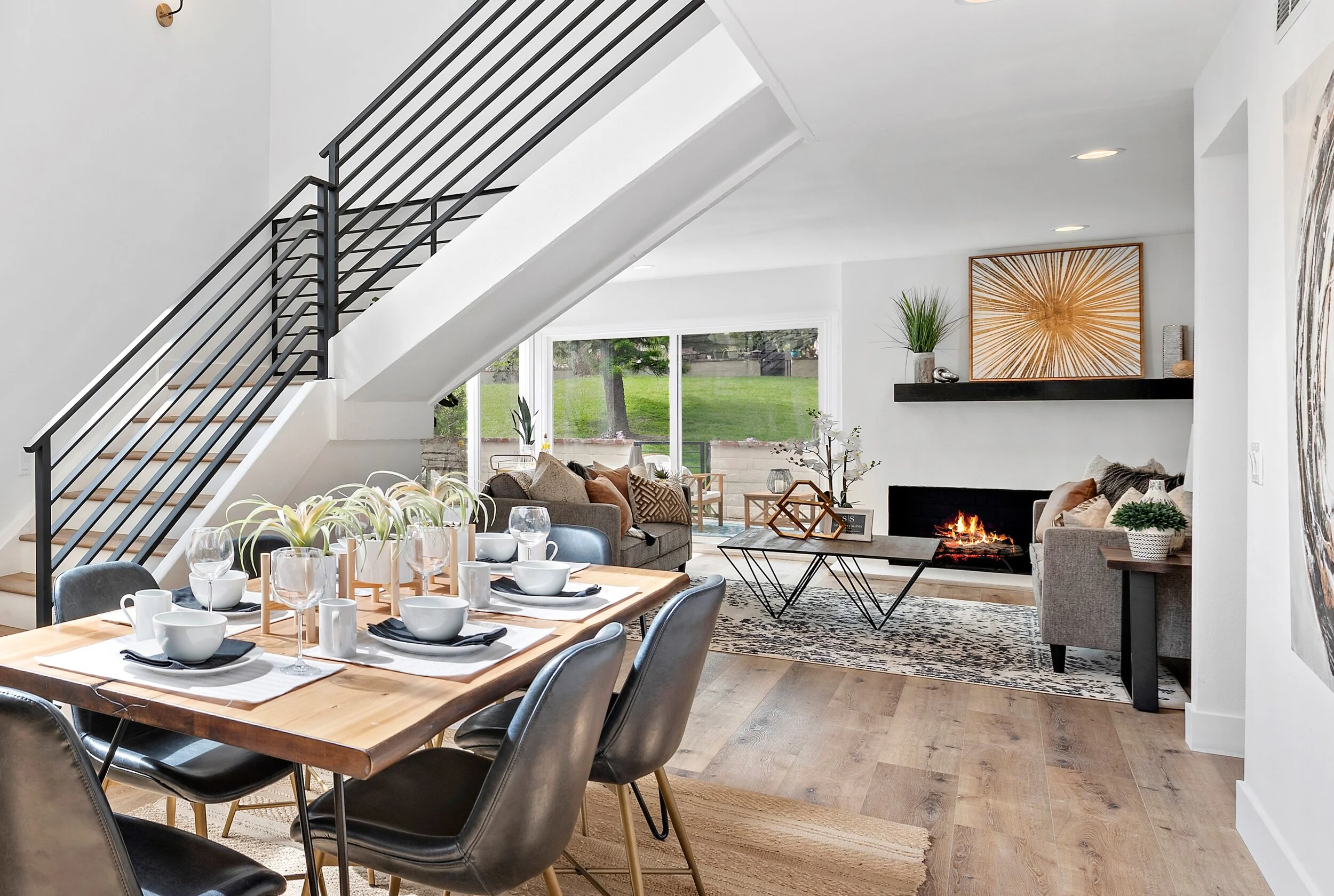 Open-concept living and dining room with staircase, fireplace, large windows, and modern decor.