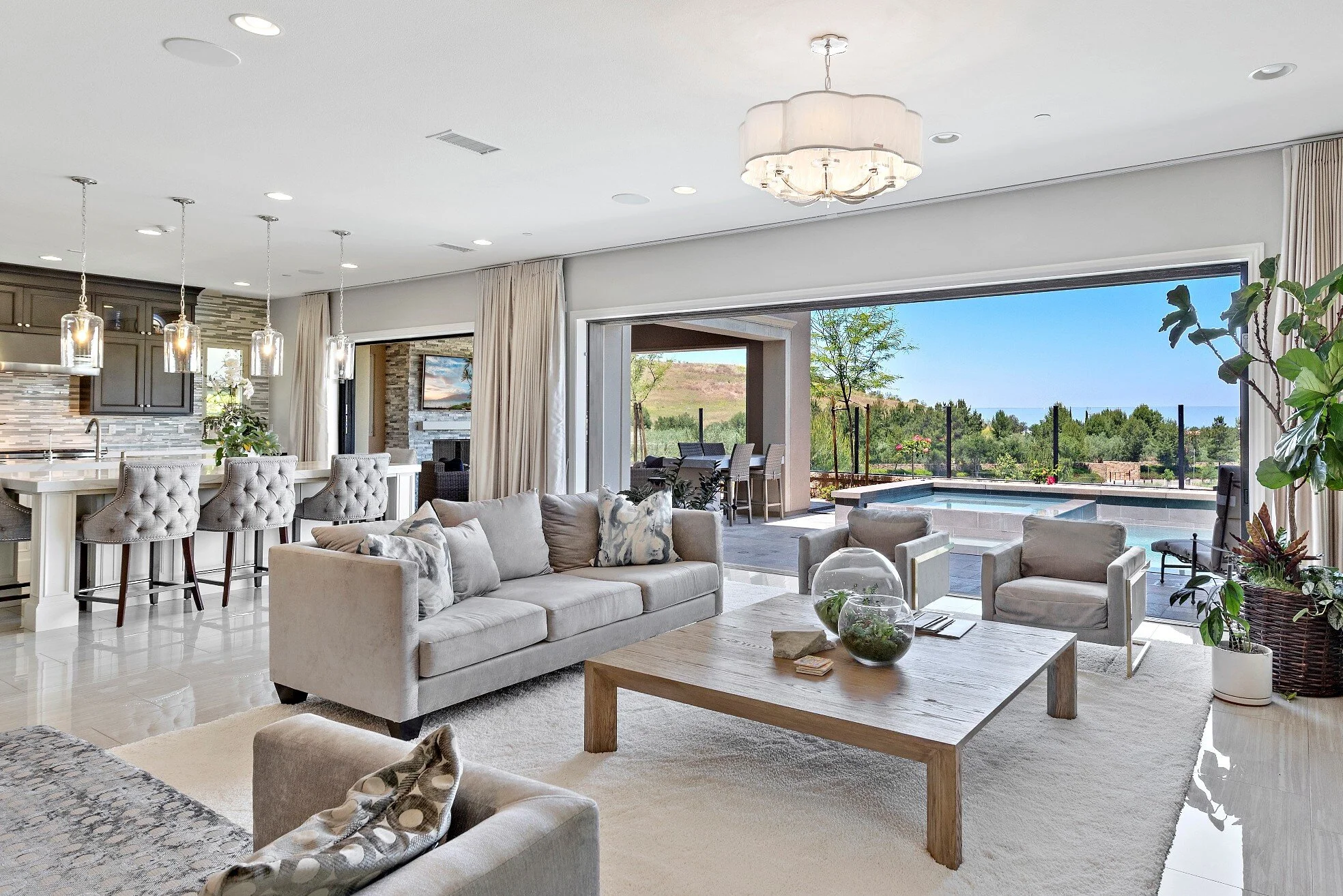 Modern living room with white sofa, armchairs, wooden coffee table, hanging pendant lights, open sliding glass door leading to outdoor patio with pool and scenic view of greenery and hills.
