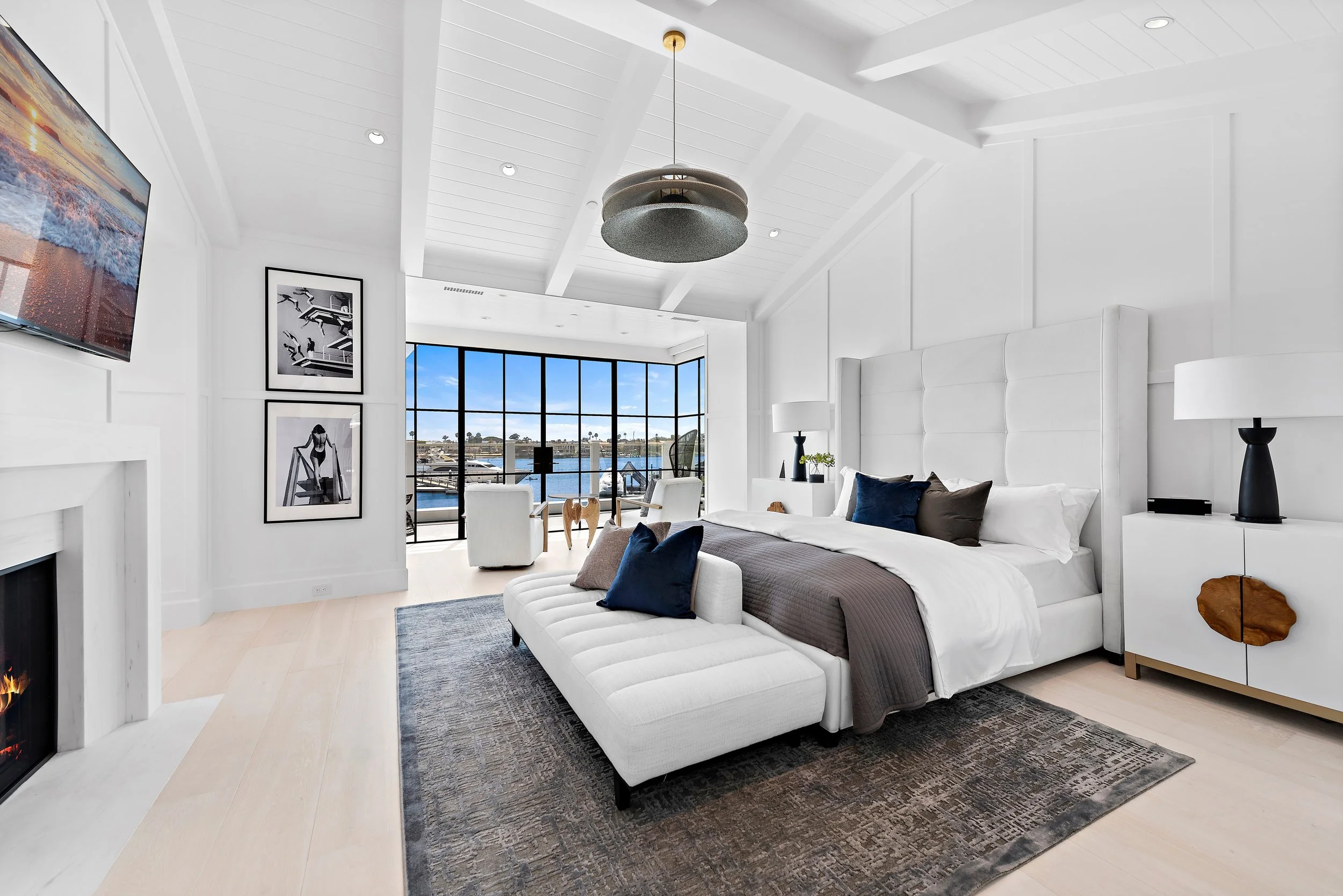 Modern bedroom with white walls, high ceiling, large window with a view of water, white furniture, and decorative pillows.