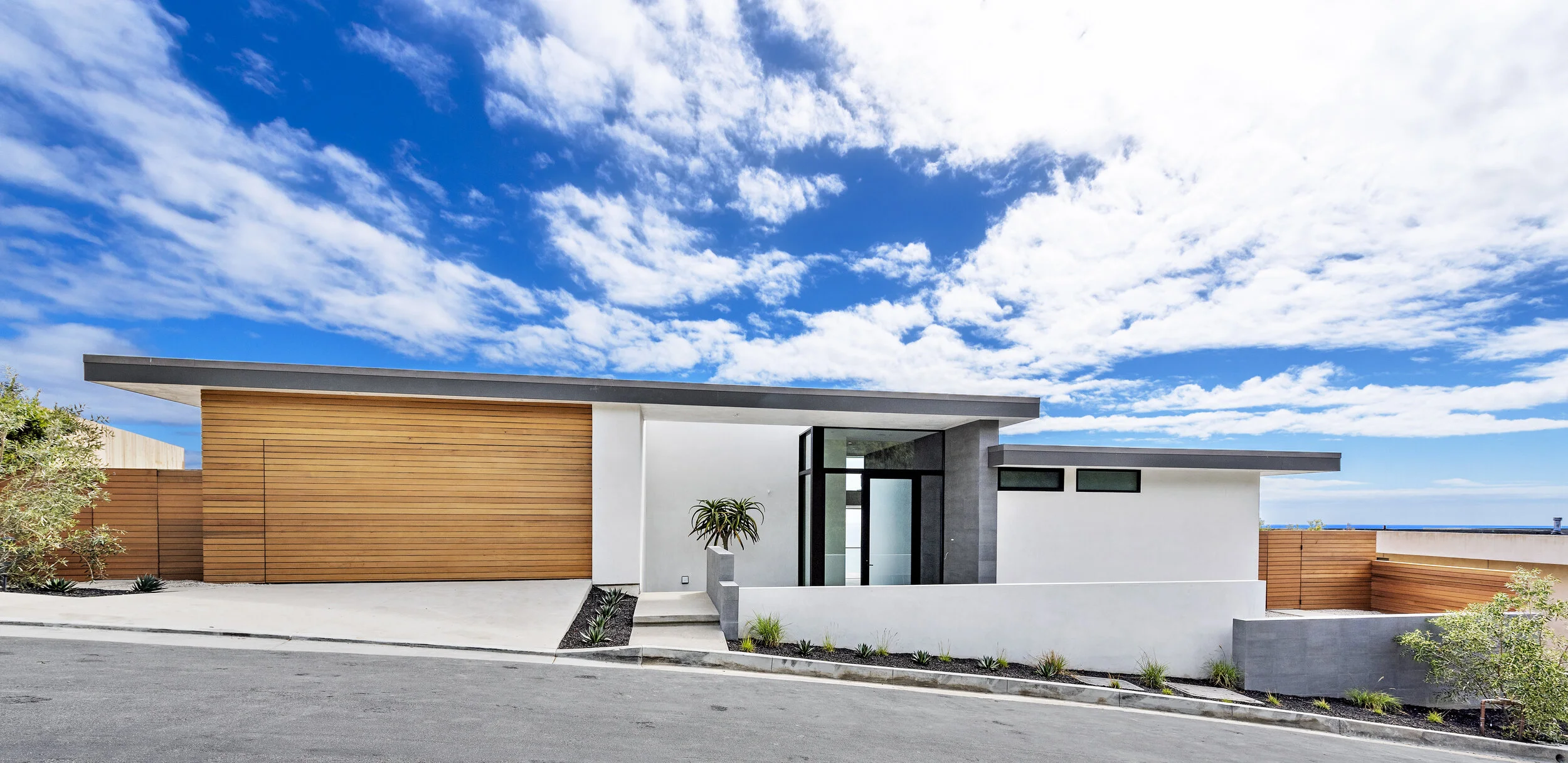 Modern house with a flat roof, white walls, gray accents, and wooden fence, under a bright blue sky with scattered clouds.