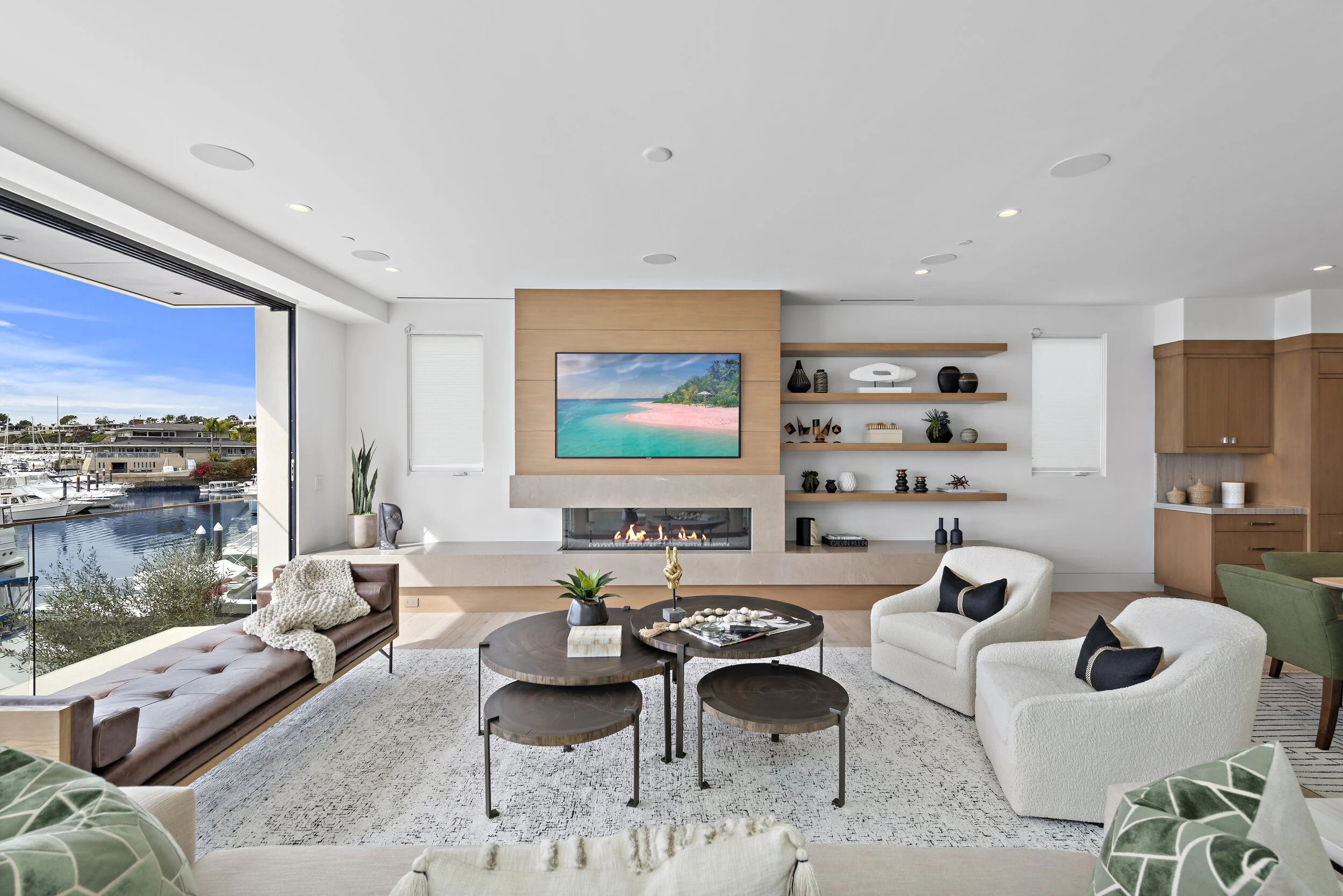 Modern living room with large window overlooking a marina, featuring a fireplace, mounted TV, white armchairs, wooden and green furniture, and decorative shelves.