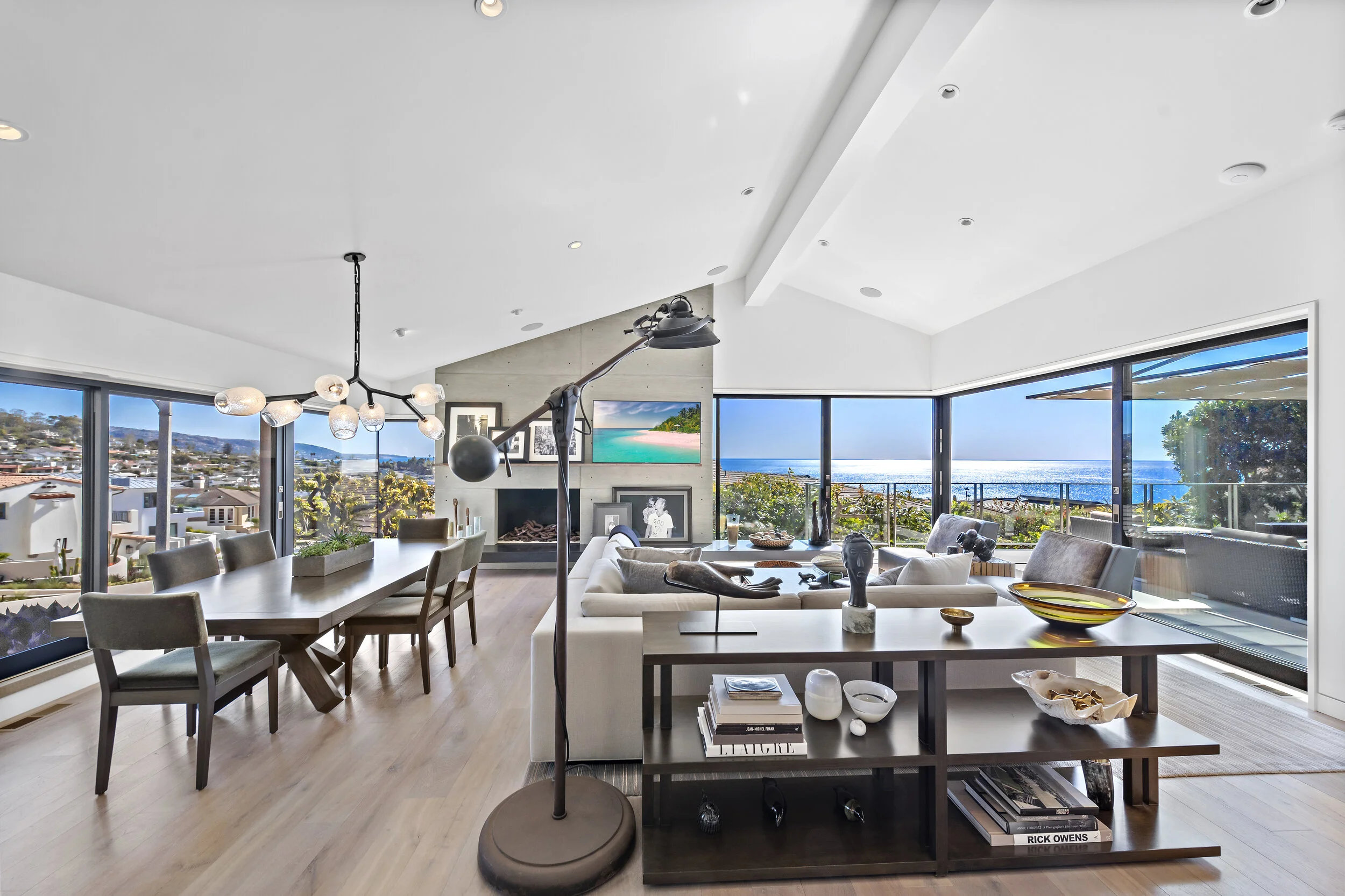 Living room with large windows overlooking a view of the ocean and a neighborhood, modern furniture including a white sectional sofa, a dining table with eight chairs, a fireplace, and decorative items on shelves and tables, with a floor lamp and con