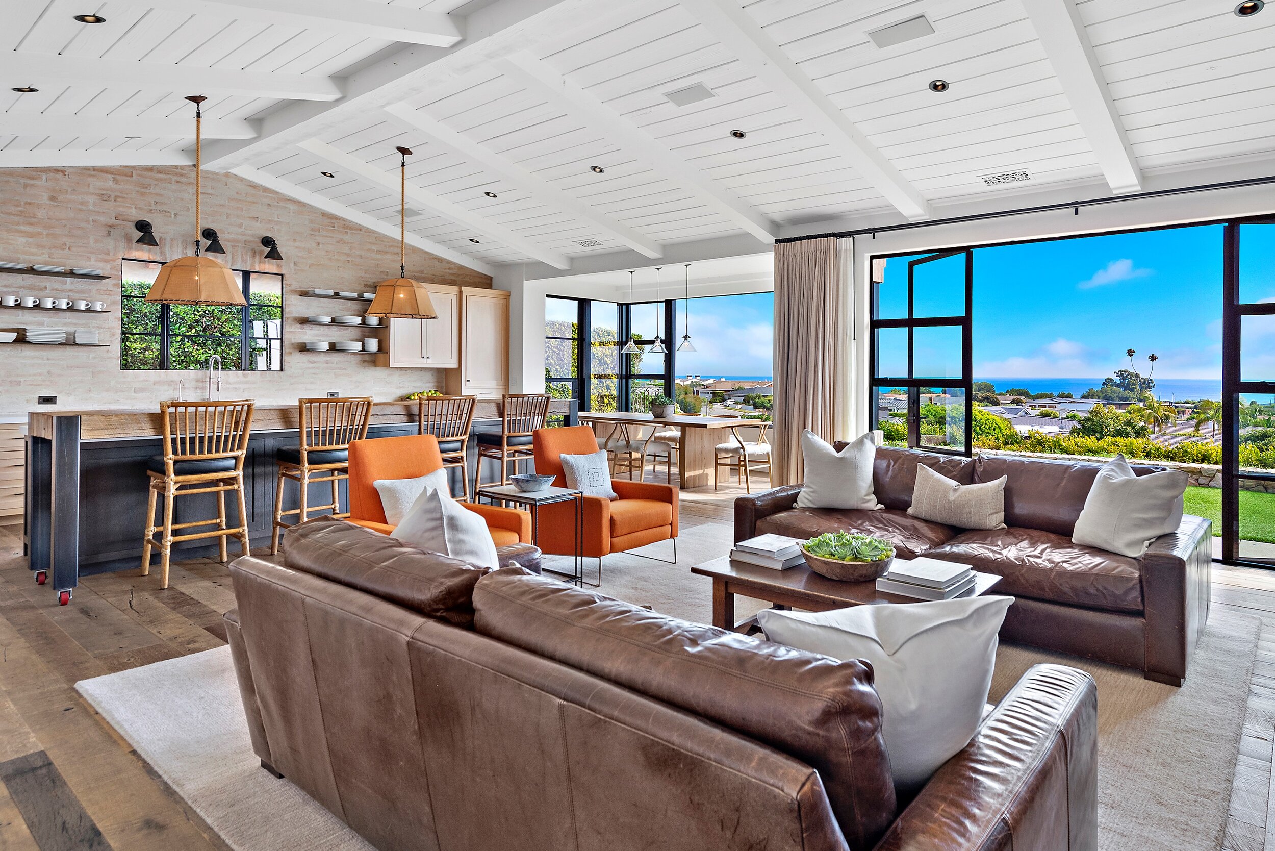 Modern open-concept living room with leather sofas, orange armchairs, large windows showing a scenic ocean view, and a kitchen area with stools and pendant lights.
