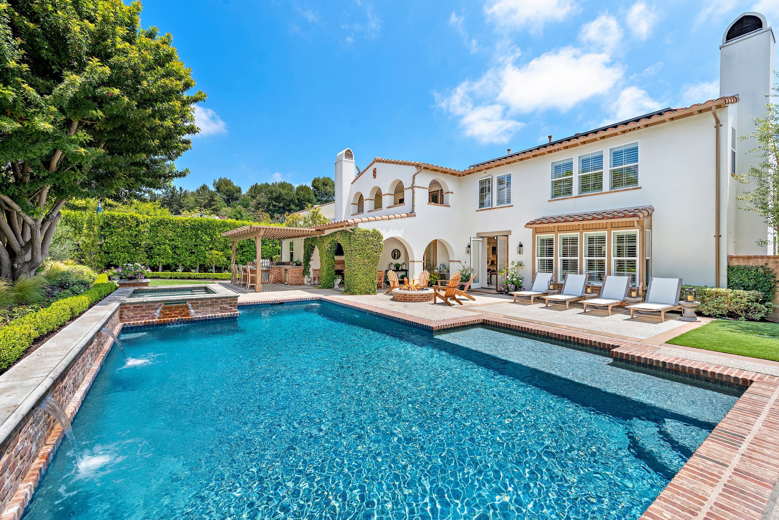 Backyard with a swimming pool, lounge chairs, outdoor dining area, lush greenery, and a white two-story house under a blue sky with clouds.