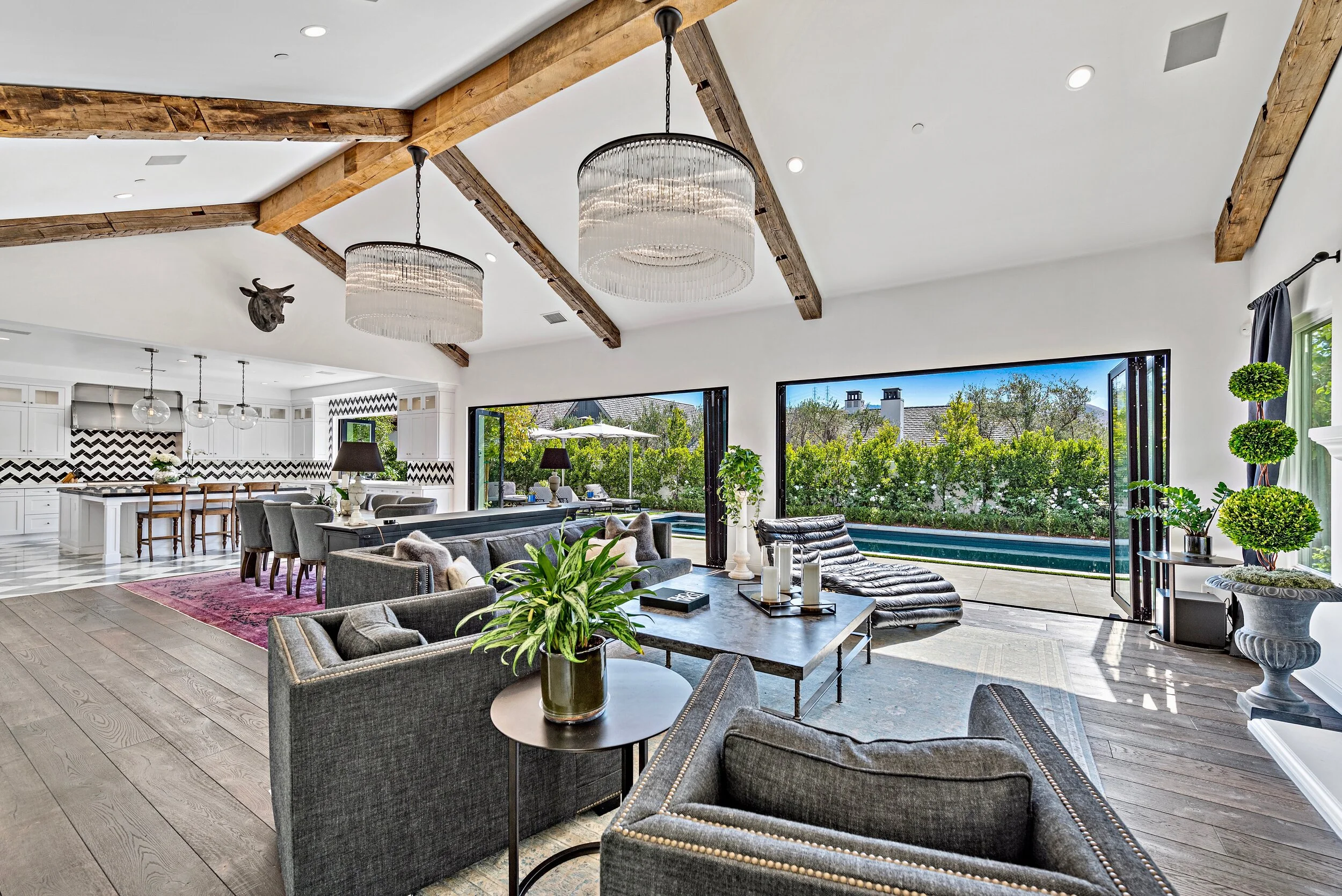Open-concept living room and kitchen with vaulted ceiling, exposed wooden beams, modern chandeliers, large windows, and sliding glass doors leading to outdoor pool area.