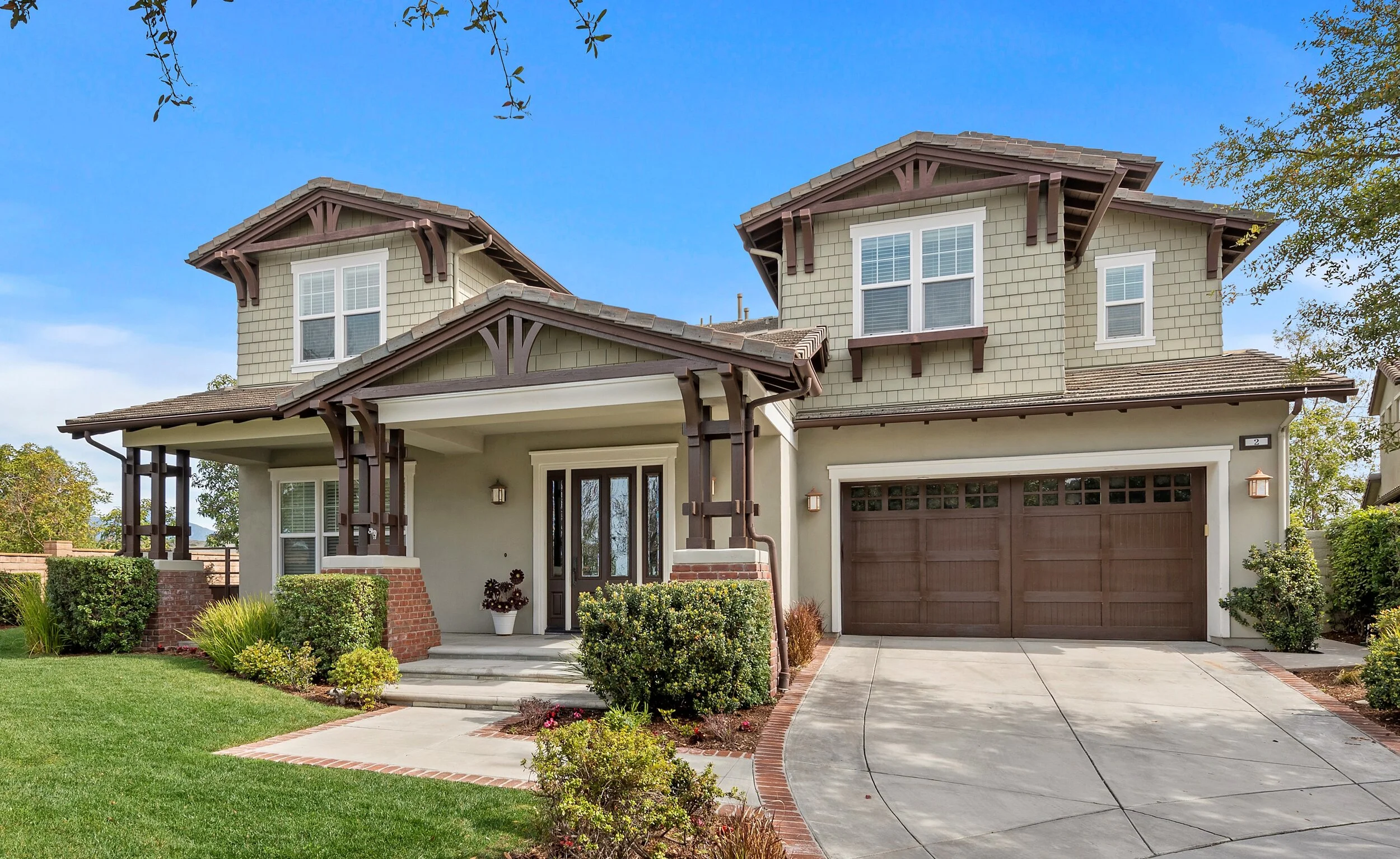 Two-story house with a front porch, brown garage door, and green shrubs in the yard, under a blue sky.