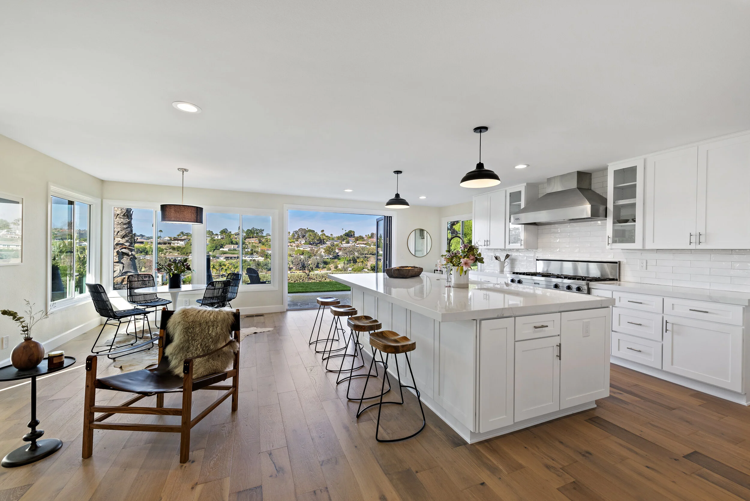 Open-concept kitchen and dining area with white cabinets, kitchen island, wooden bar stools, and large windows overlooking a green landscape and blue sky.
