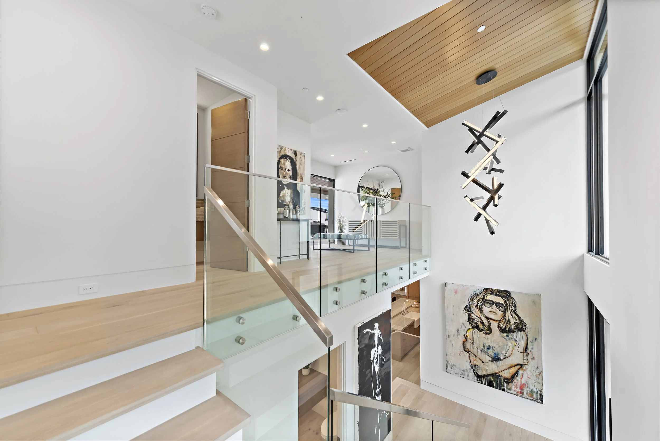 Modern loft-style interior with wooden and white walls, glass railing, contemporary art paintings, a round mirror, and a sculptural black and white chandelier.