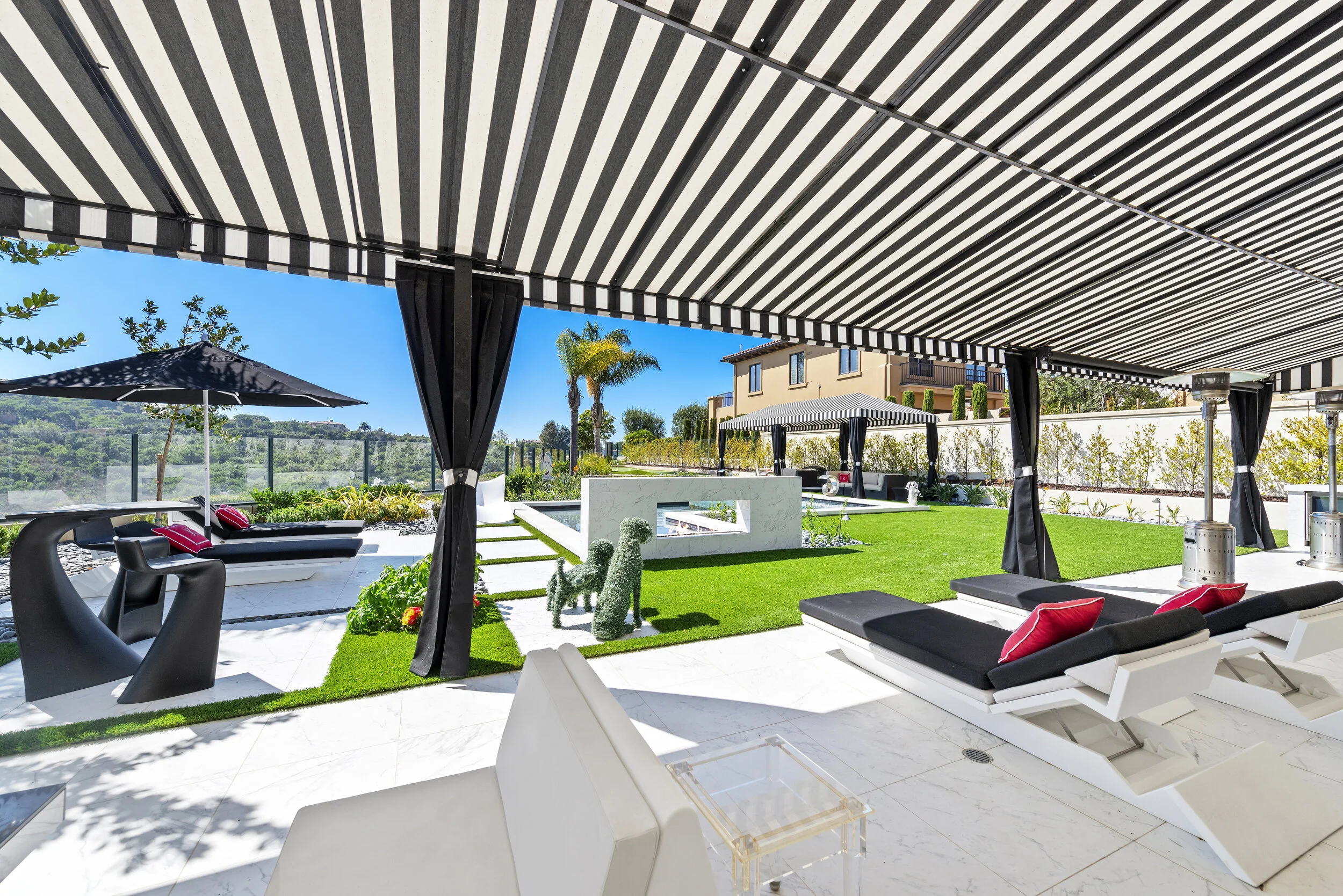 Outdoor luxury lounge area with black and white striped awning, lounge chairs with red pillows, black sun chairs, greenery, and a white modern building in the background.