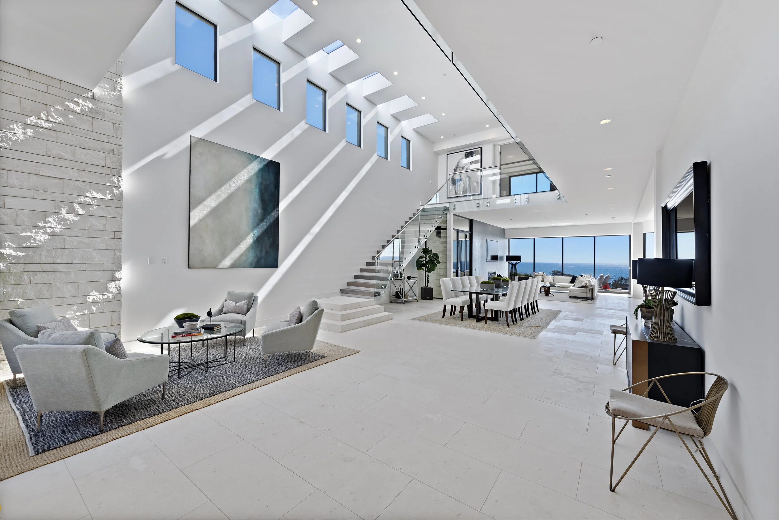 Modern spacious living room with white walls, large windows with ocean views, high ceiling, and modern furniture including a white sofa and armchairs, a dining table with white chairs, and minimal decor.
